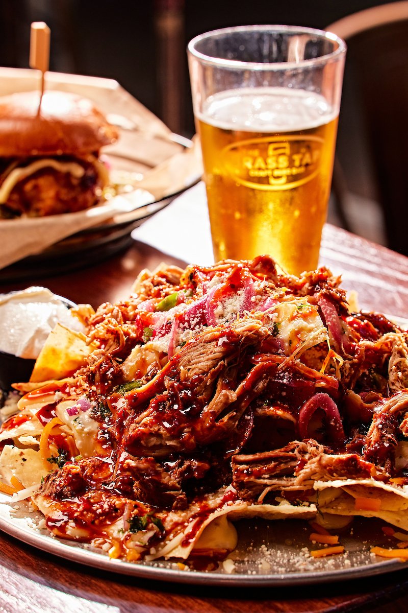 A plate of shredded beef topped with barbecue sauce, served with cheese, vegetables, and a side of sour cream, with a glass of beer and a burger in the background.