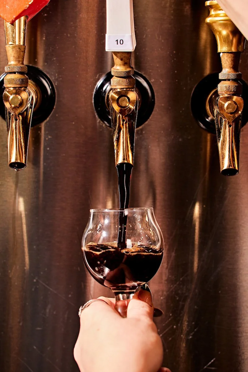 A person filling a glass with draft dark beer from a tap in a bar or brewery setting.