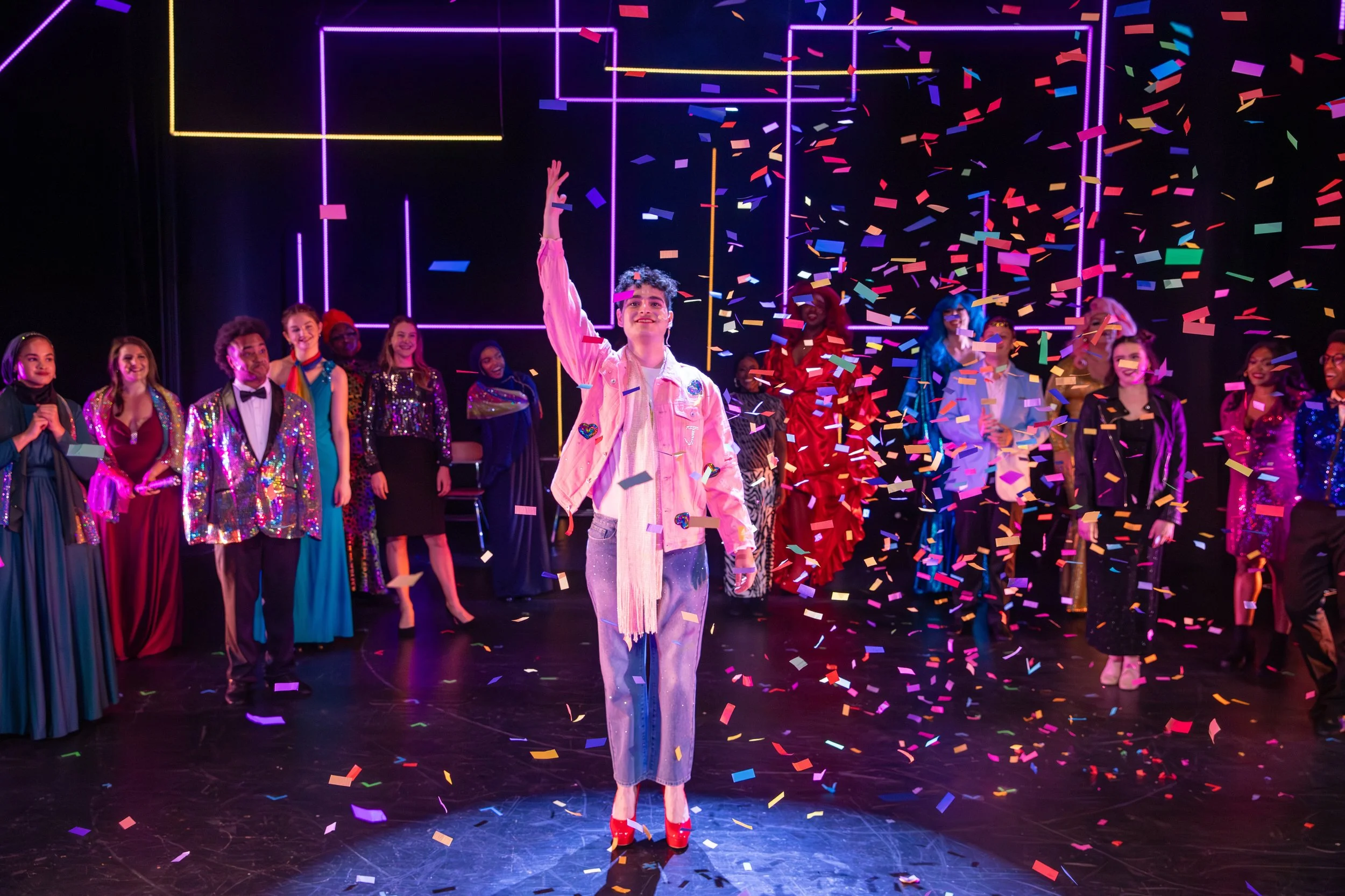 A group of diverse performers dressed in colorful costumes, standing on stage with confetti falling around them, celebrating a performance or event.