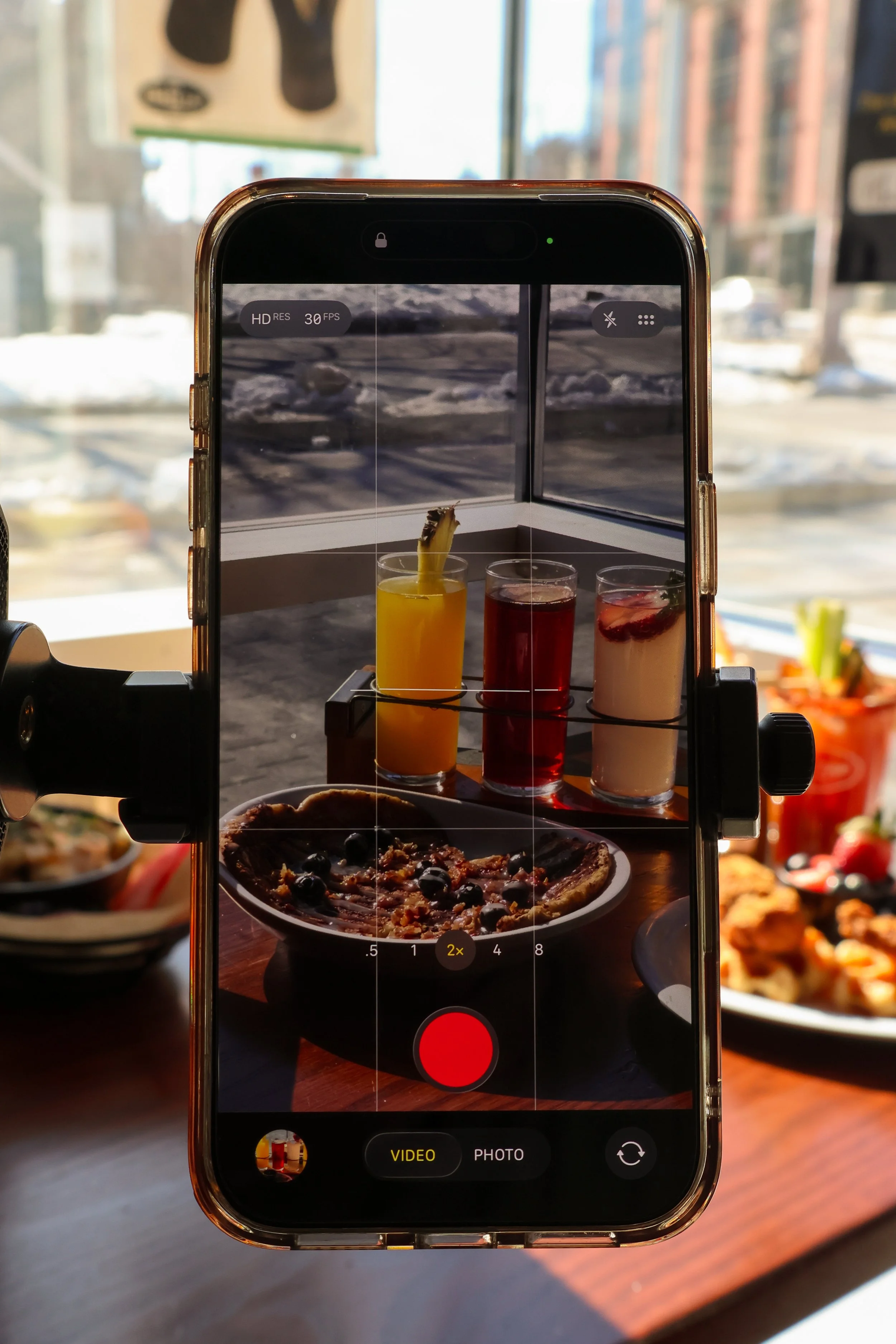 A smartphone on a tripod captures a photo of three drinks and a plate of flapjacks in a restaurant. The drinks are colorful, with one garnished with a pineapple slice and fruit slices, while the flapjacks have blueberries on top.