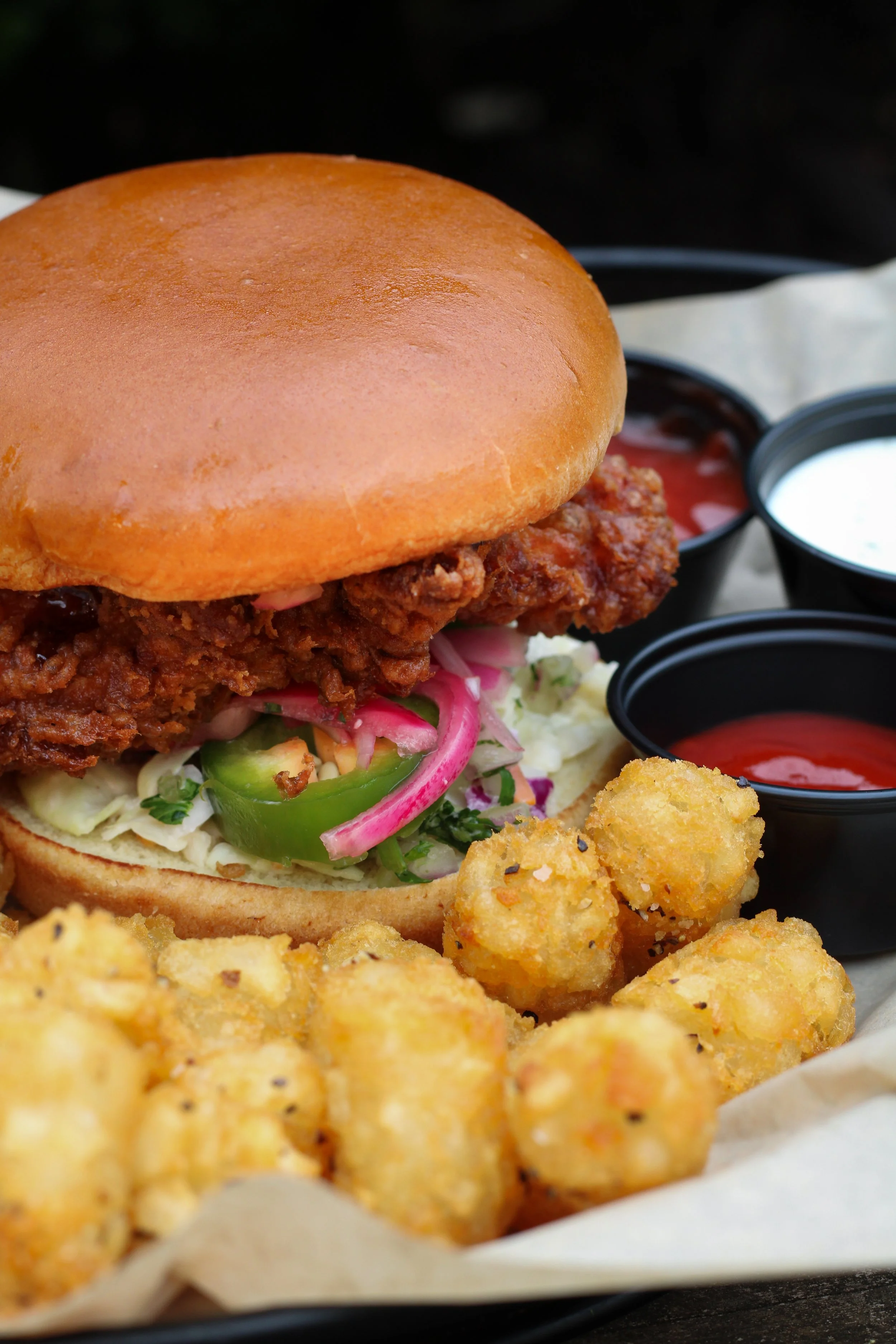 Close-up of a fried chicken sandwich with pickles, onions, and sauce on a bun, with fried tots and dipping sauces.