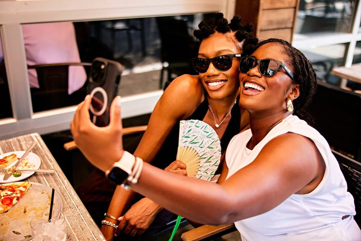 Two women sitting at a table, smiling and taking a selfie together, wearing sunglasses, with pizza on the table.