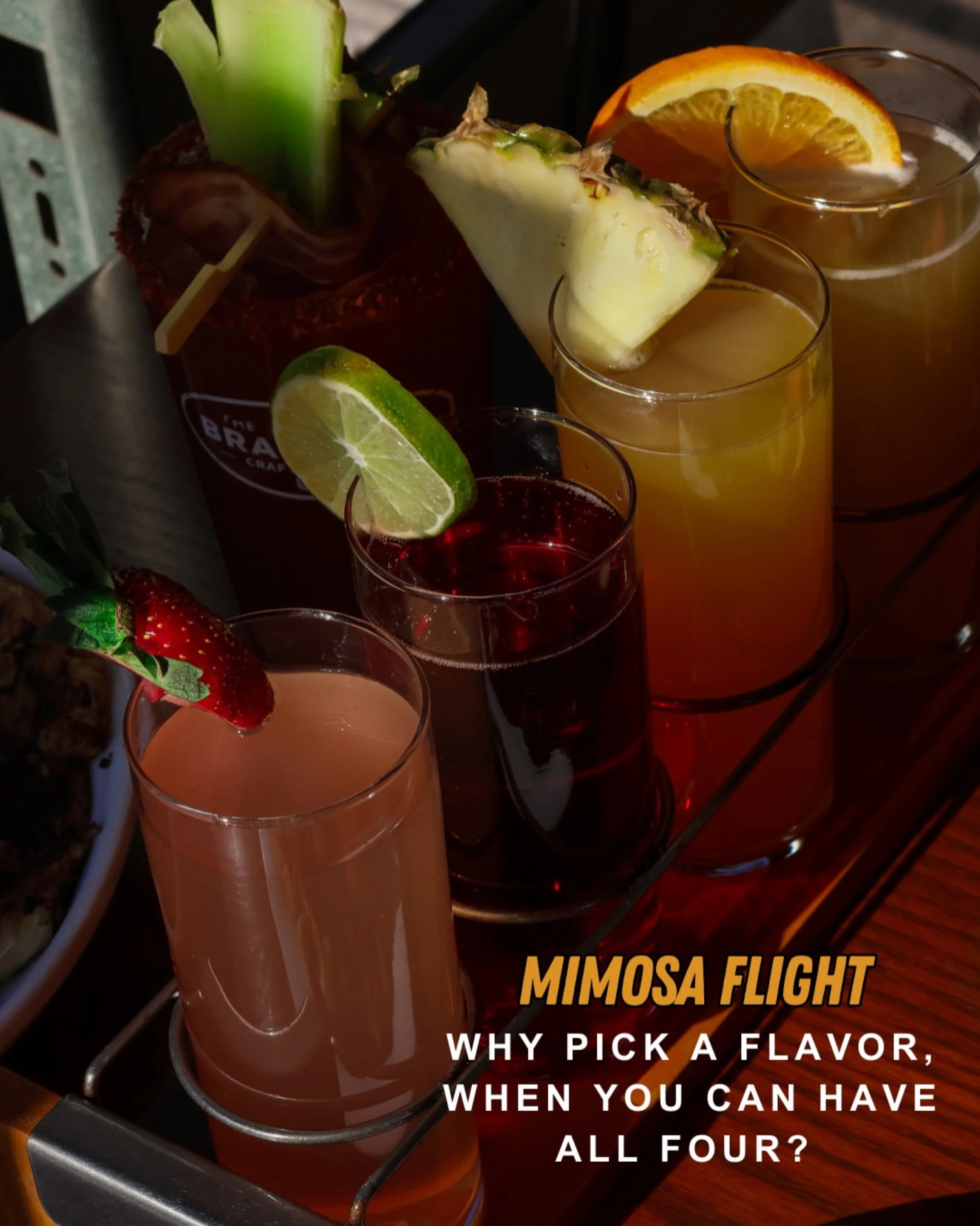 A tray of four colorful drinks with garnishes including orange slice, pineapple, lime, and strawberry, arranged on a metal rack.