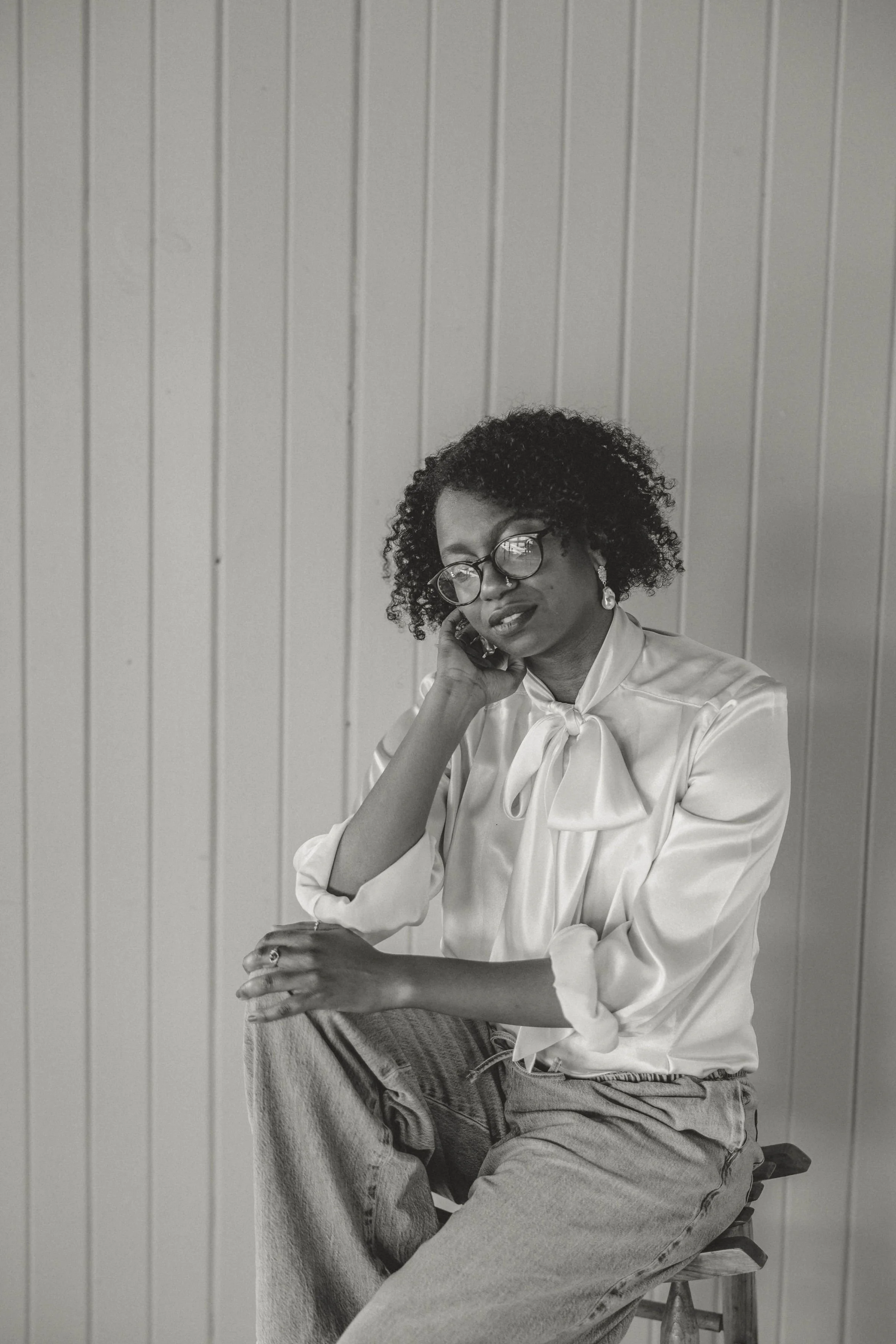 Black and white photo of a black woman with curly hair wearing glasses, a satin blouse with a bow, and earrings. She is sitting on a stool against a panel wall, with her hand resting on her neck and her other hand on her knee, looking thoughtful.