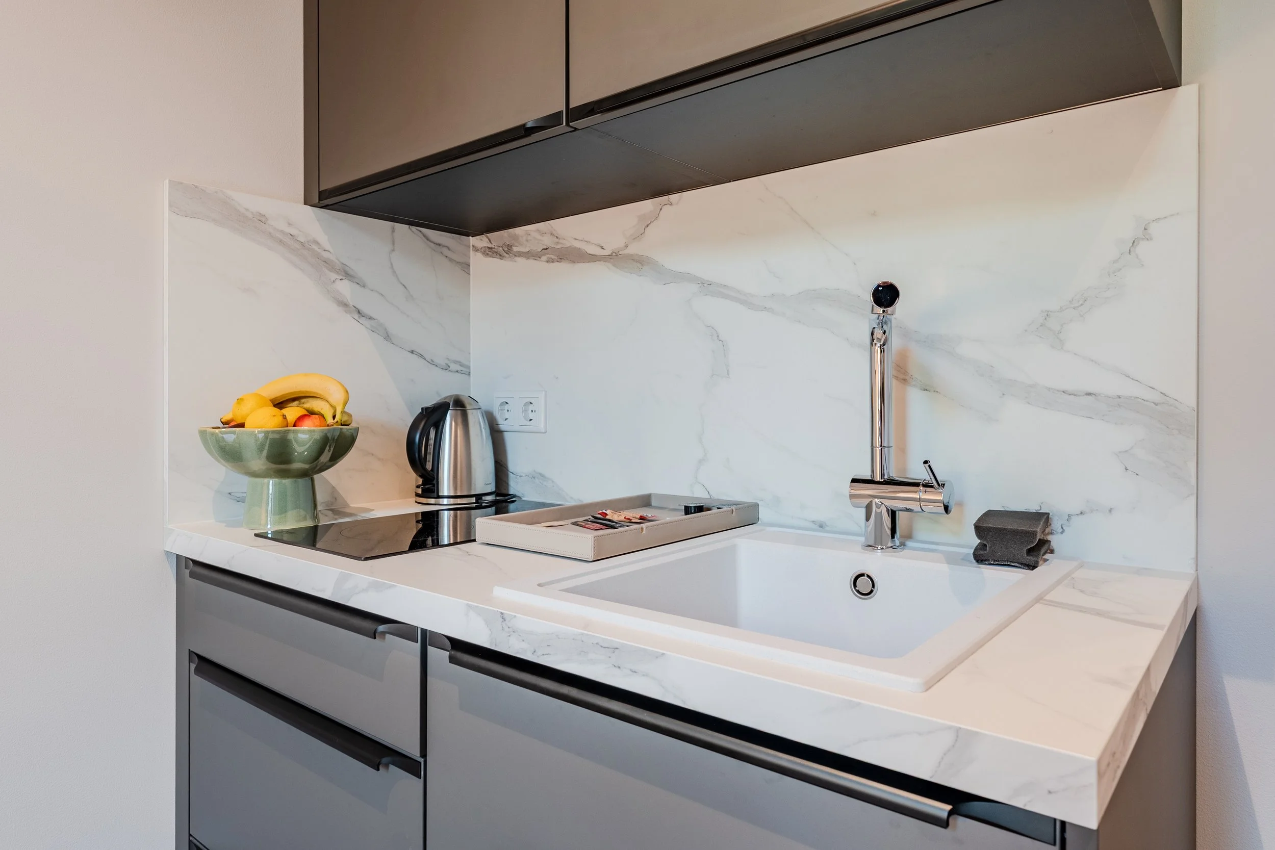 Modern kitchen with white marble backsplash and countertop, black cabinets, and a sink with a chrome faucet. A bowl of assorted fruit sits on the counter, alongside a stainless steel electric kettle and a tray with small packets.