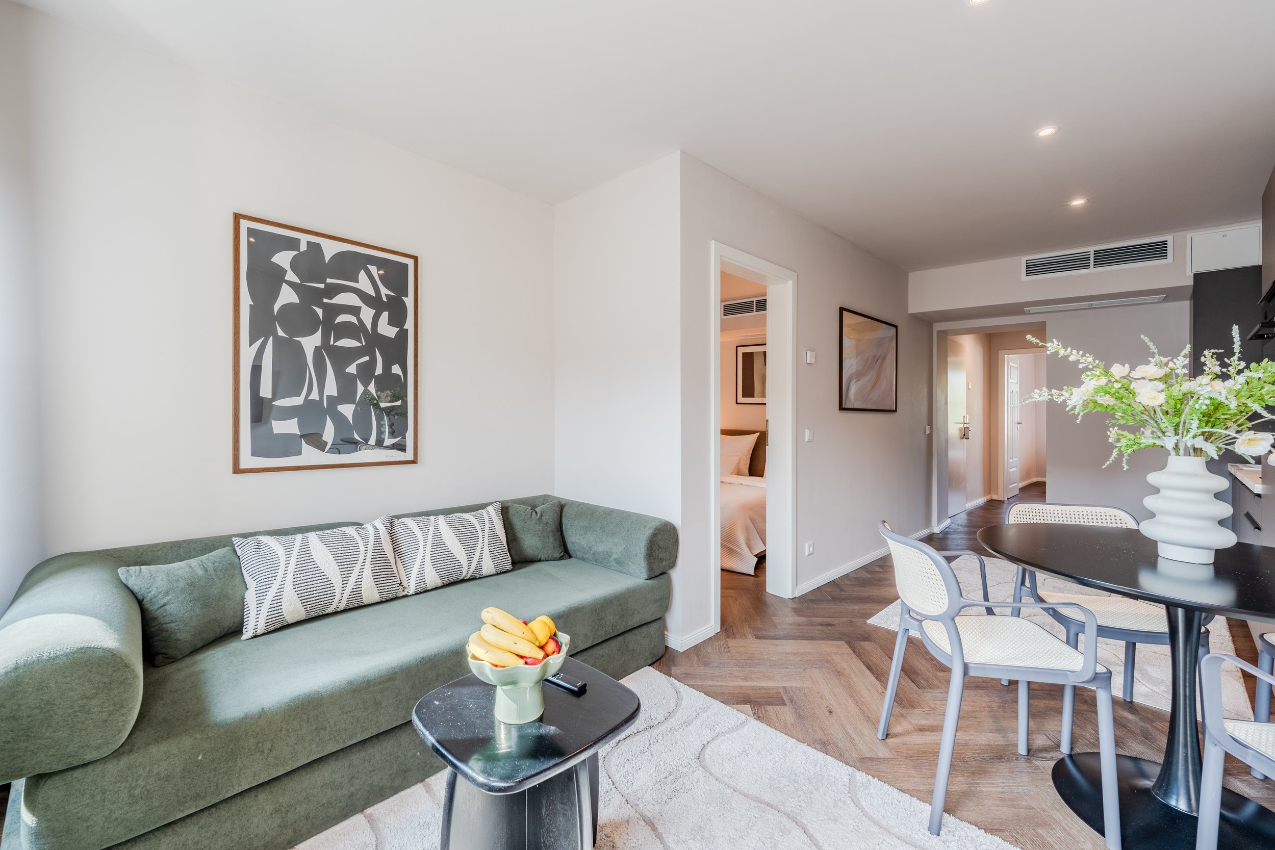 Living room with green sofa, abstract wall art, wooden flooring, dining area with white chairs, large vase with white flowers, bowl of fruit, doorway to bedroom.