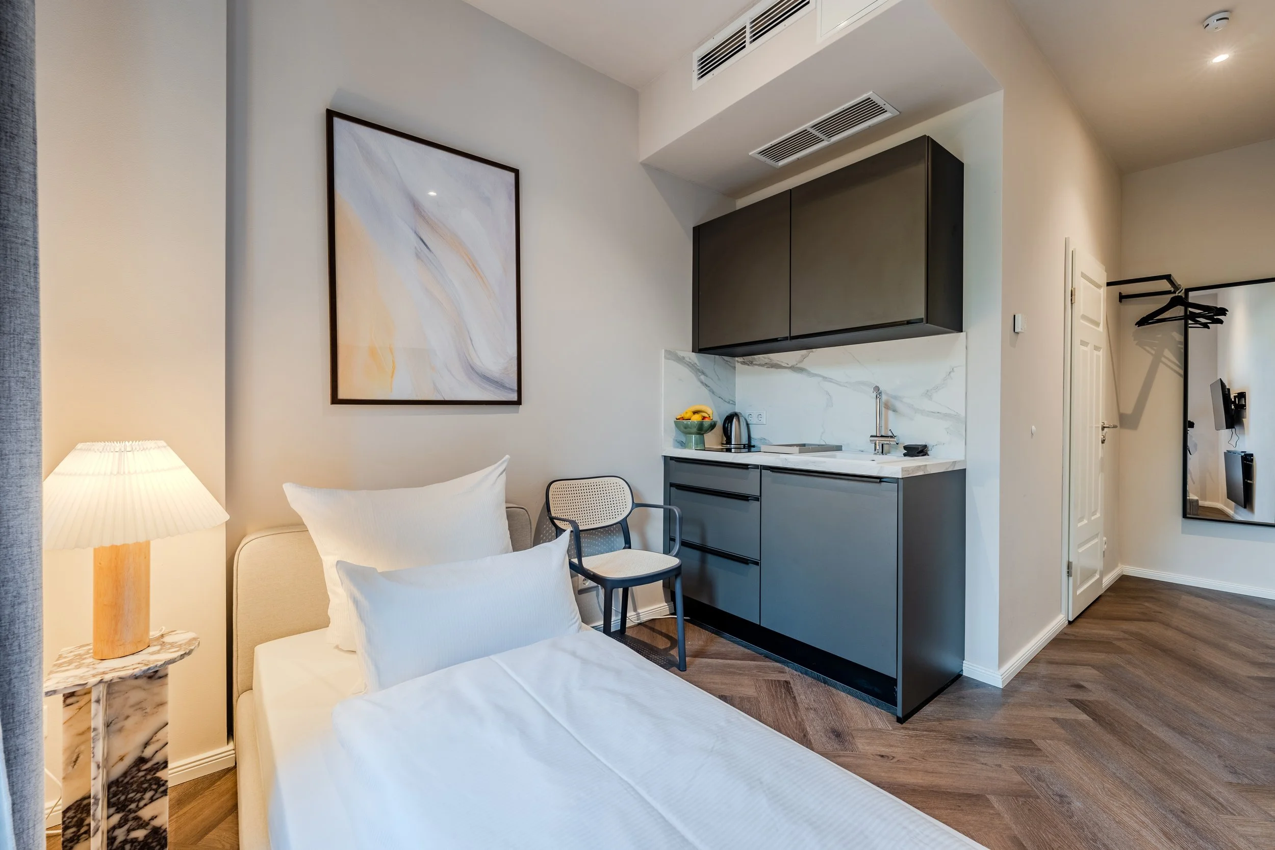 Interior of a modern studio apartment with a bed, a side table, artwork on the wall, a kitchenette with dark cabinets, marble backsplash, and a small dining chair.
