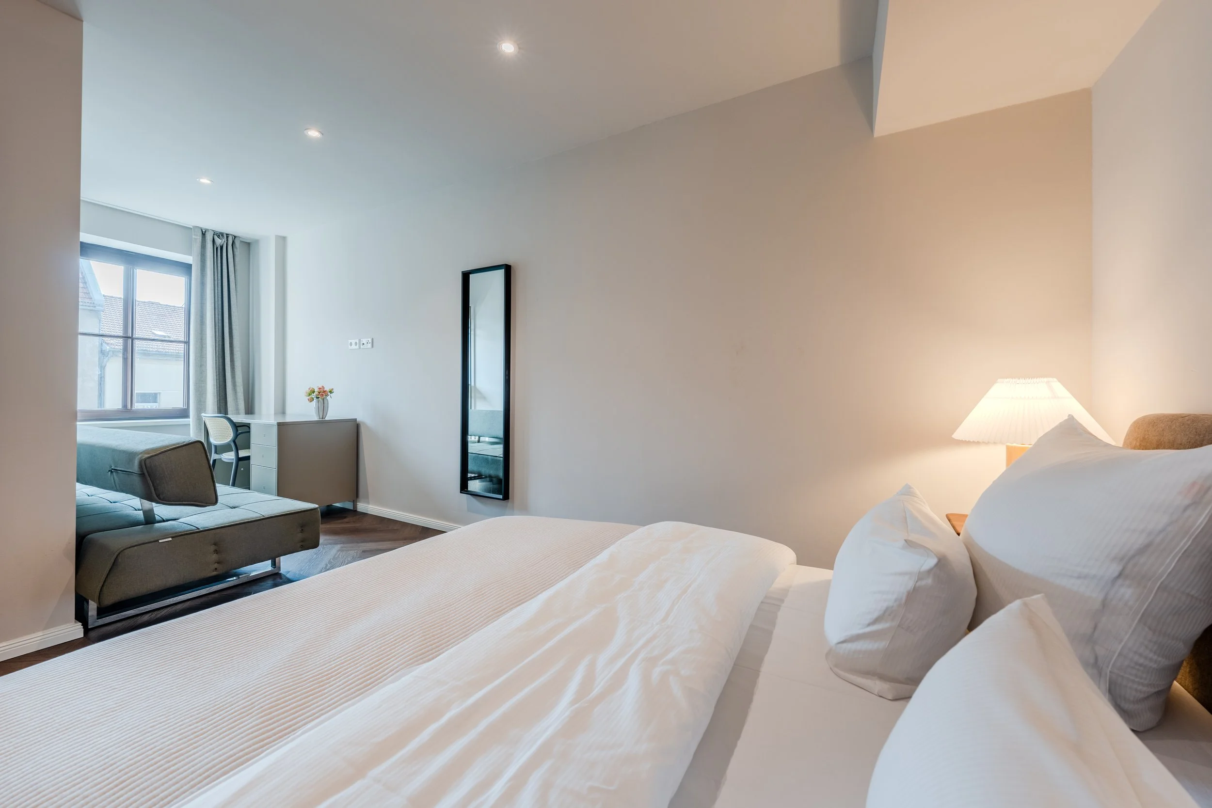 A modern hotel room with a bed, nightstand, lamp, mirror, desk, and chair near a window with curtains.