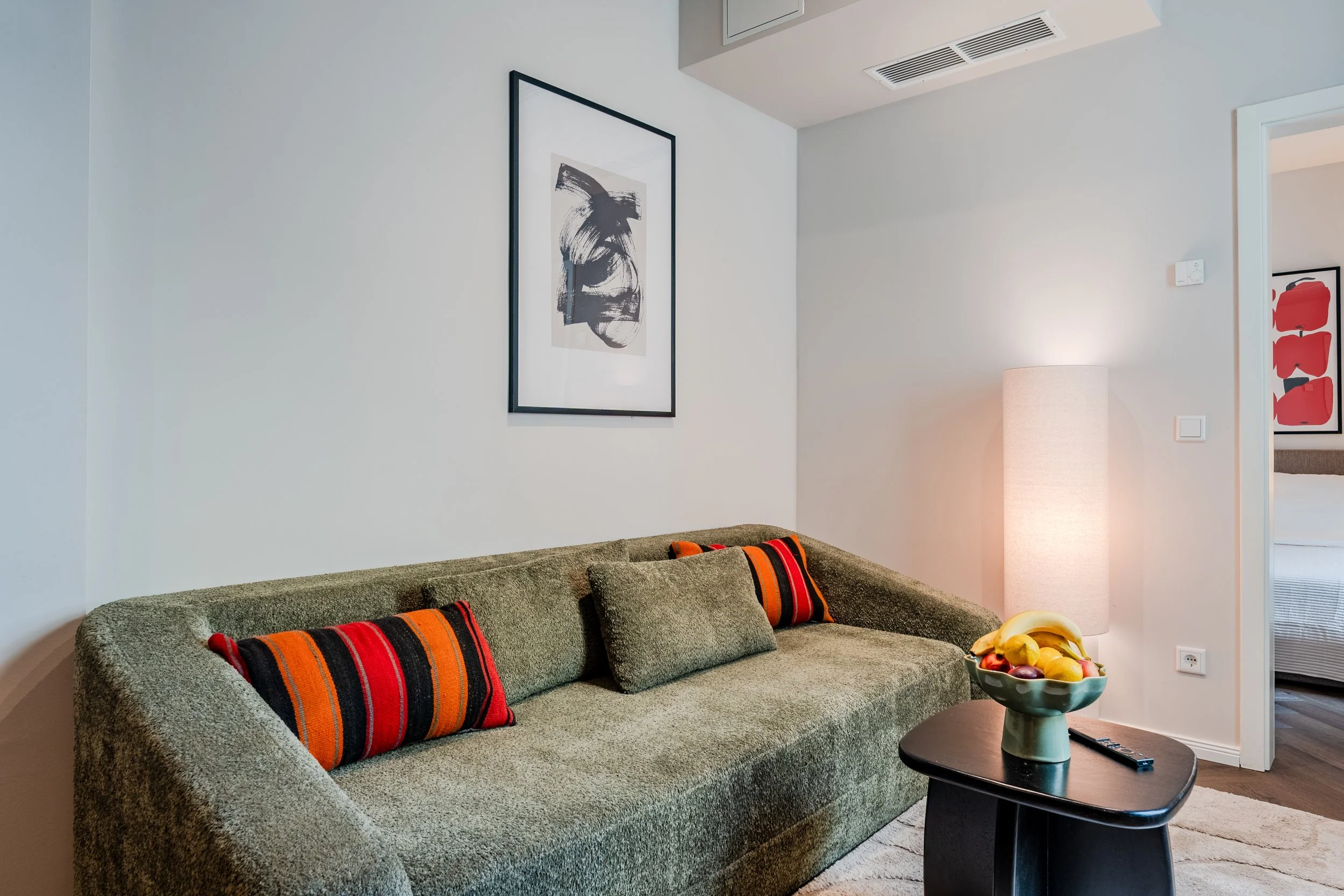 A living room with a textured gray sofa decorated with striped throw pillows in red, orange, and black. A black coffee table holds a bowl of fruit and a remote control. On the wall, there are two art pieces: a black and white abstract print and a colorful piece with red, black, and white shapes. A tall, white floor lamp illuminates the space, and a doorway to the right reveals part of another room with similar decor.