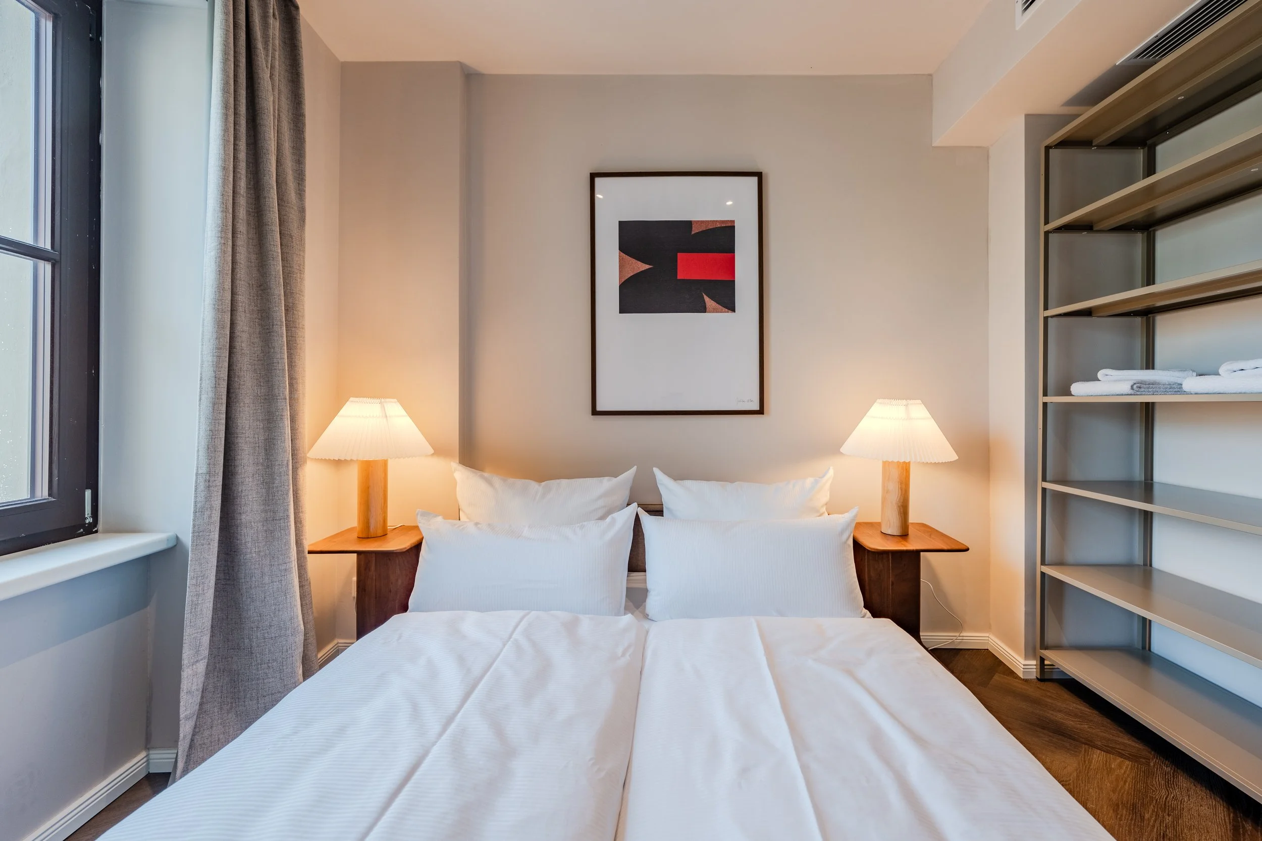 A cozy bedroom with a large bed featuring white linens and four pillows. There are two wooden nightstands with matching lamps on each side of the bed. A framed abstract art piece hangs above the headboard. To the right, open shelving holds neatly folded white towels. A window with beige curtains is on the left, and a section of an air vent is visible on the ceiling.