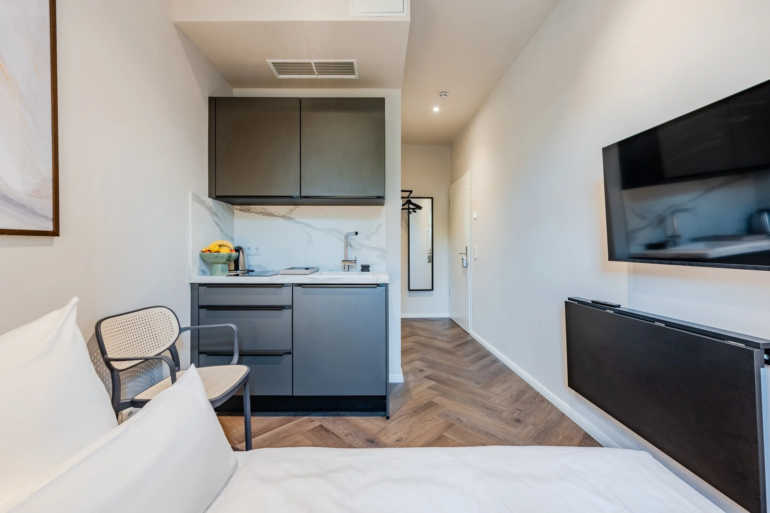 Interior of a modern apartment with a small kitchen area, white walls, a wall-mounted TV, a dining chair, and a couch with pillows, with a hallway leading to a door.