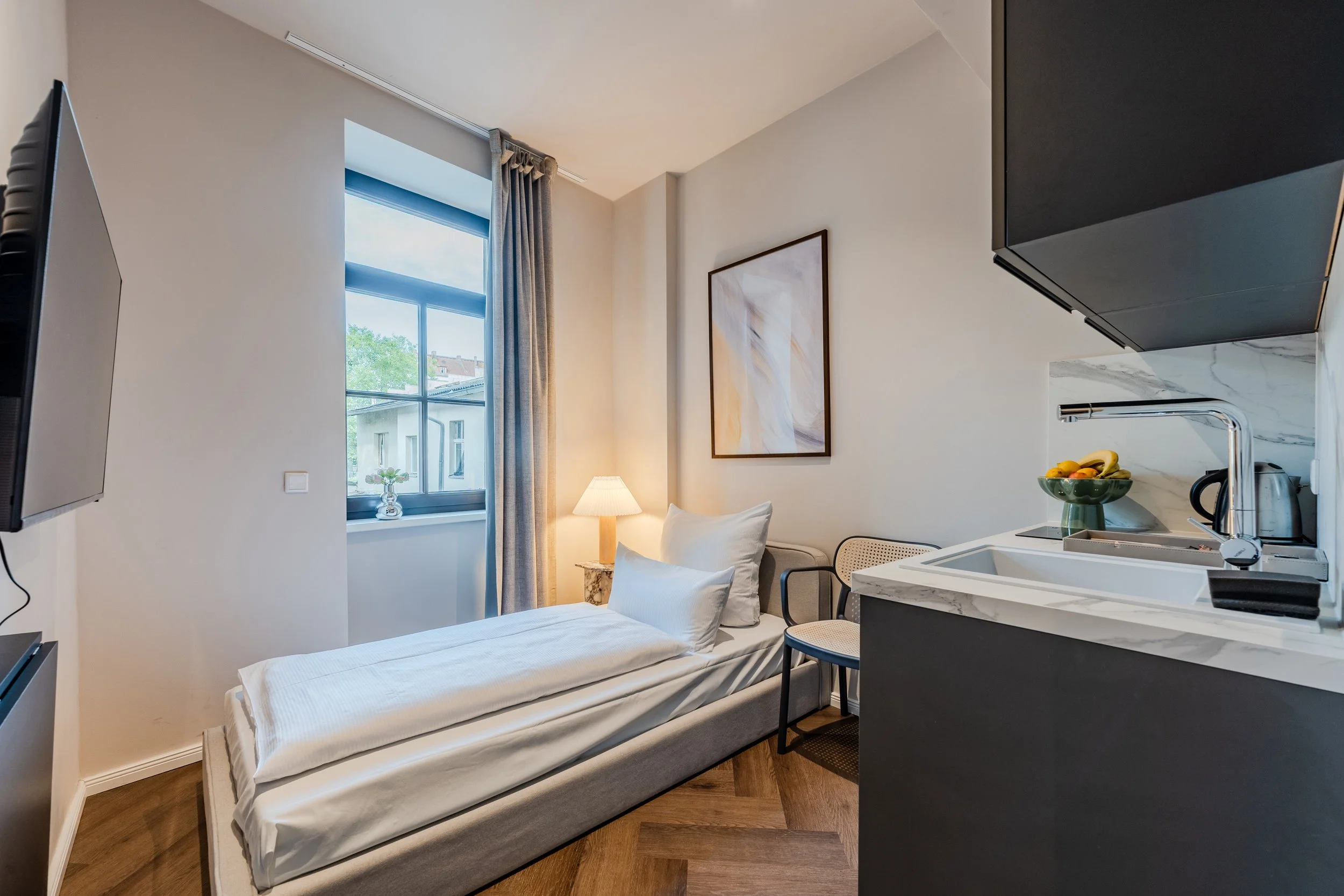 Interior view of a small modern studio apartment featuring a bed, a window with curtains, a painting on the wall, a small side table with a lamp, and a kitchenette with a bowl of fruit.