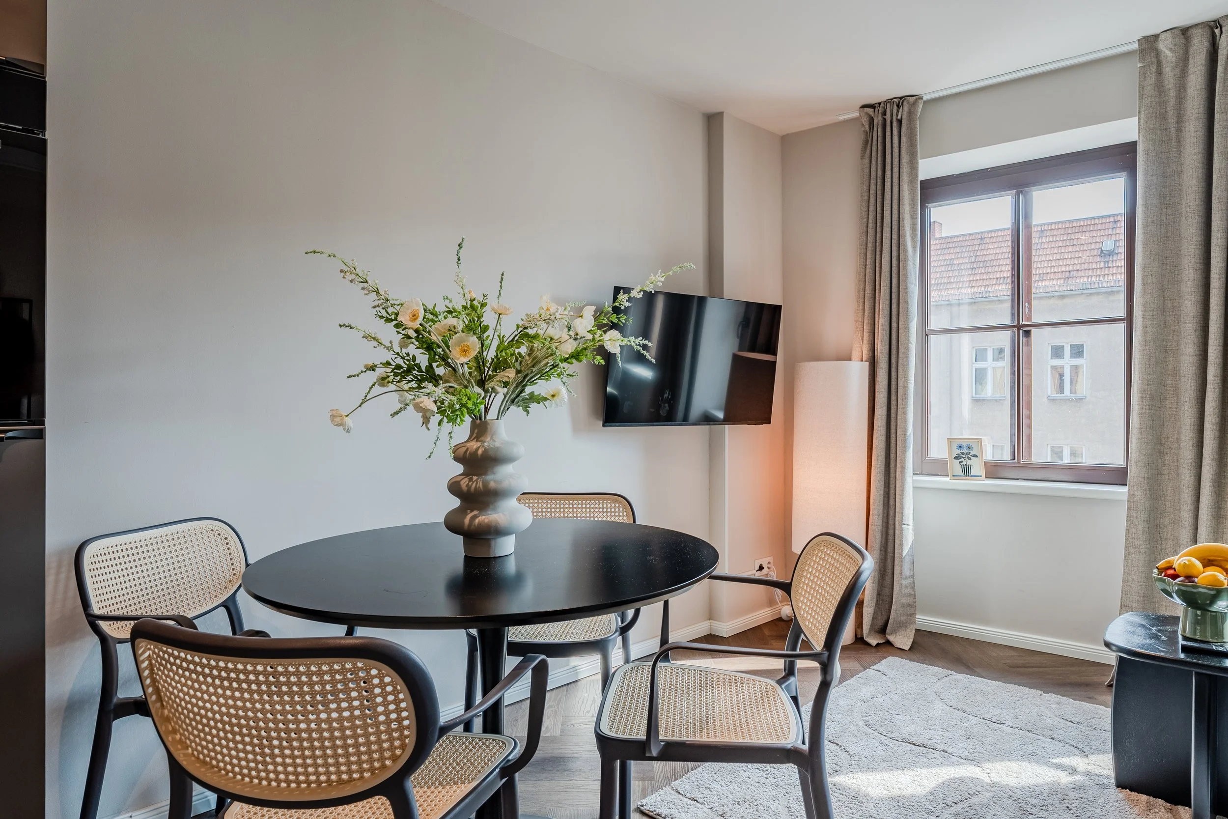 A dining area with a black round table, beige rattan chairs, a white vase with flowers, a flat-screen TV on the wall, a window with curtains, and a framed picture on the windowsill.