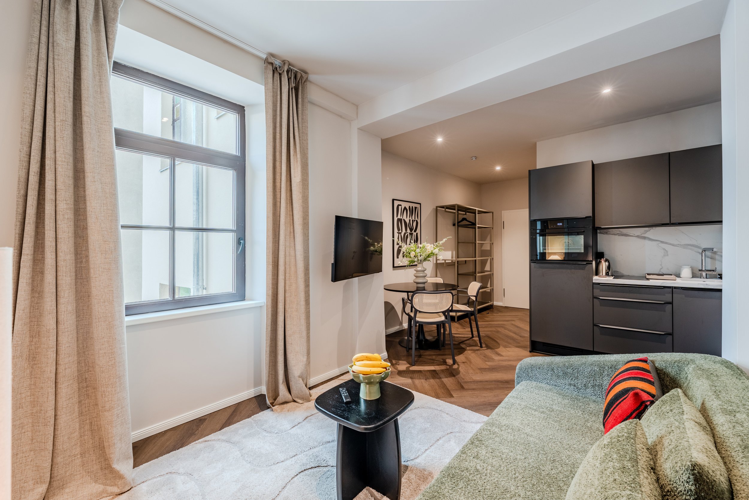Living room with large window and beige curtains, a TV on the wall, a small round dining table with chairs, kitchen area with black cabinets, marble backsplash, and coffee station, green sofa with striped pillow, black coffee table with bananas, and a rug.