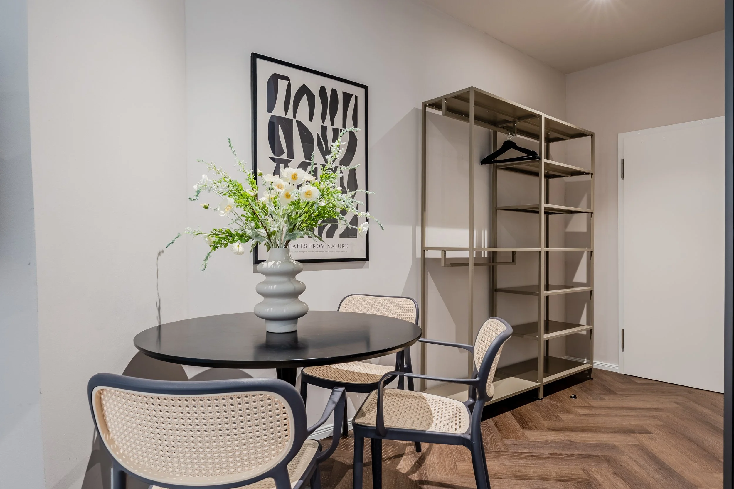 A round black table with a white vase of white flowers, surrounded by beige and black chairs, in a modern room with white walls and a metal shelving unit with clothing hangers.