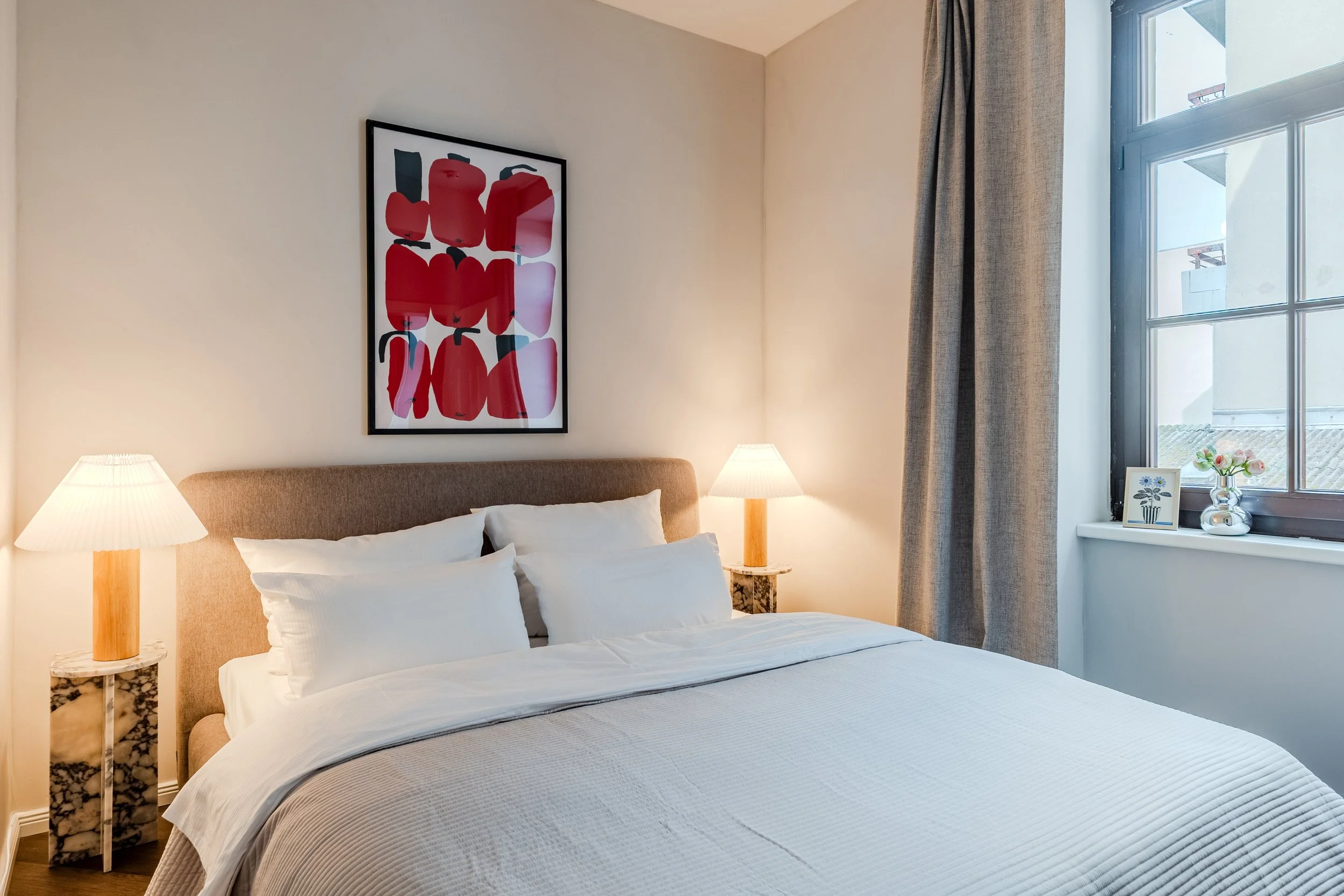 A neatly made bed with white pillows and bedding in a modern hotel room. Two matching bedside tables with marble bases and wooden lamps are on either side of the bed. A framed abstract artwork with red and black shapes hangs above the bed. A window with beige curtains and a windowsill with decorative items such as a framed picture and a vase of flowers are visible to the right.