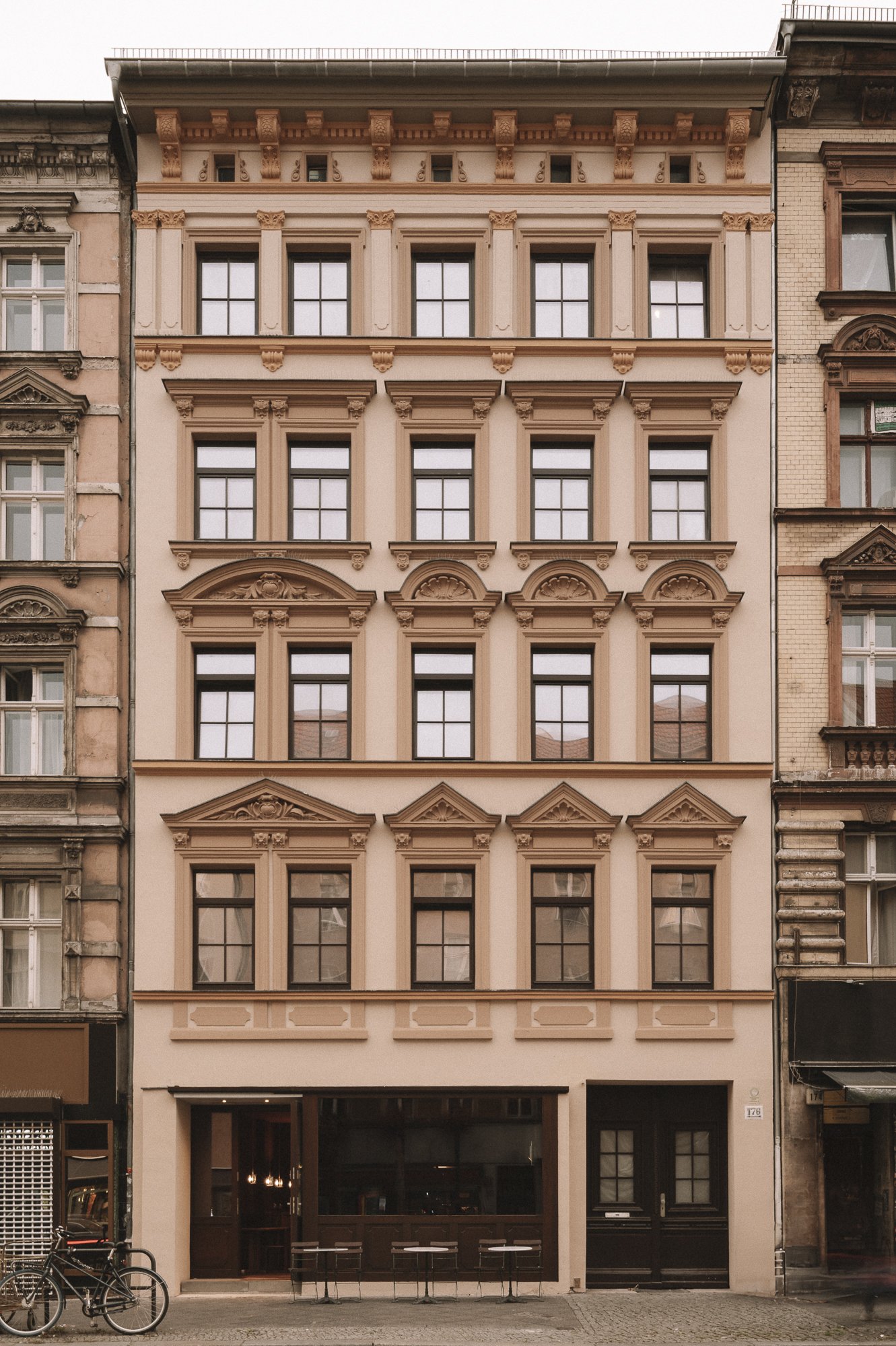 A pink classic European-style building with five floors, ornate window frames, and architectural details, positioned between neighboring buildings on a city street.