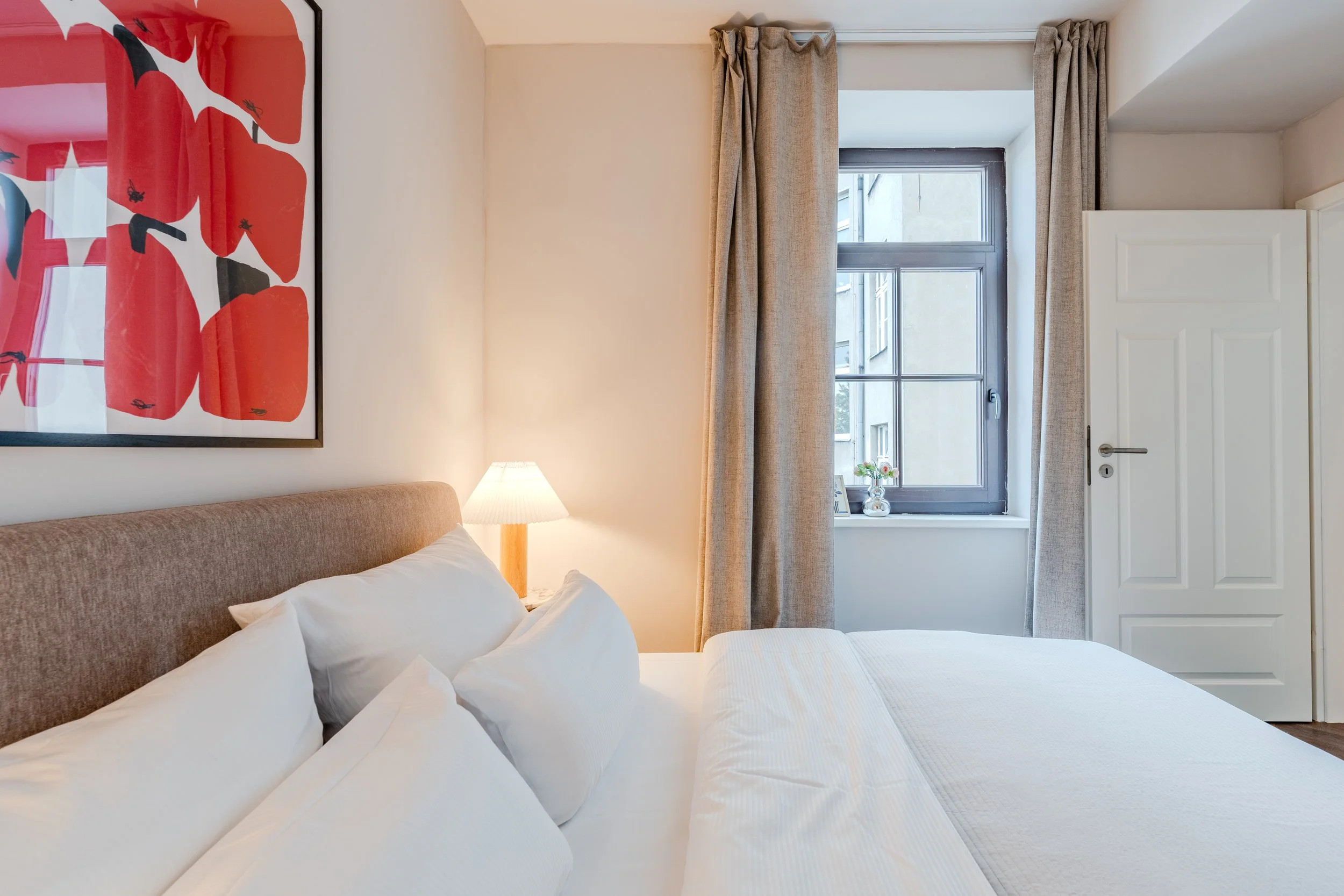 A cozy bedroom with a bed featuring white linens and pillows, a beige upholstered headboard, a bedside lamp, and a large colorful abstract art piece on the wall. There is a window with beige curtains and a white wardrobe near the door.