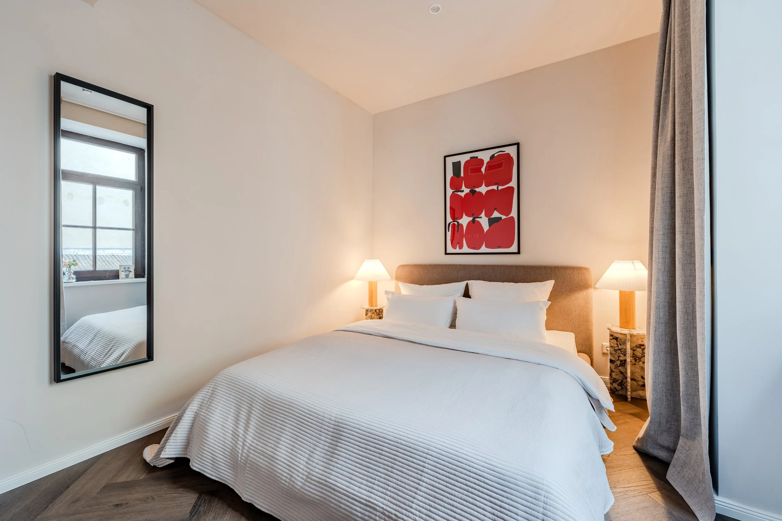 A bedroom with a large bed dressed in white bedding, two bedside lamps, an abstract red and black artwork above the bed, a tall mirror on the left, a window with gray curtains on the right, and wooden flooring.