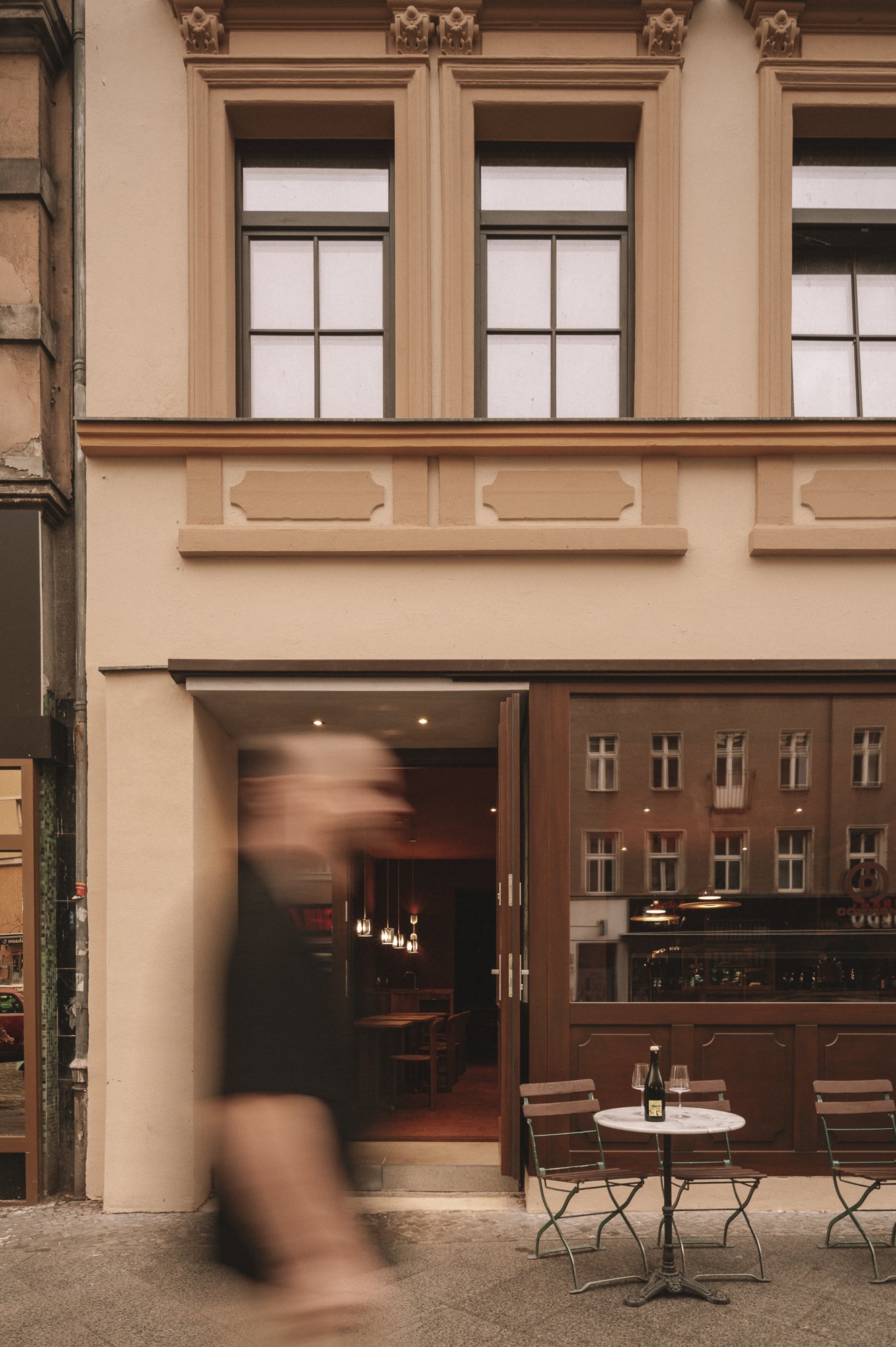A blurred person walking past a cafe with a small outdoor table and chairs, a bottle and glass on the table, in front of a building with large windows and ornate architectural details.