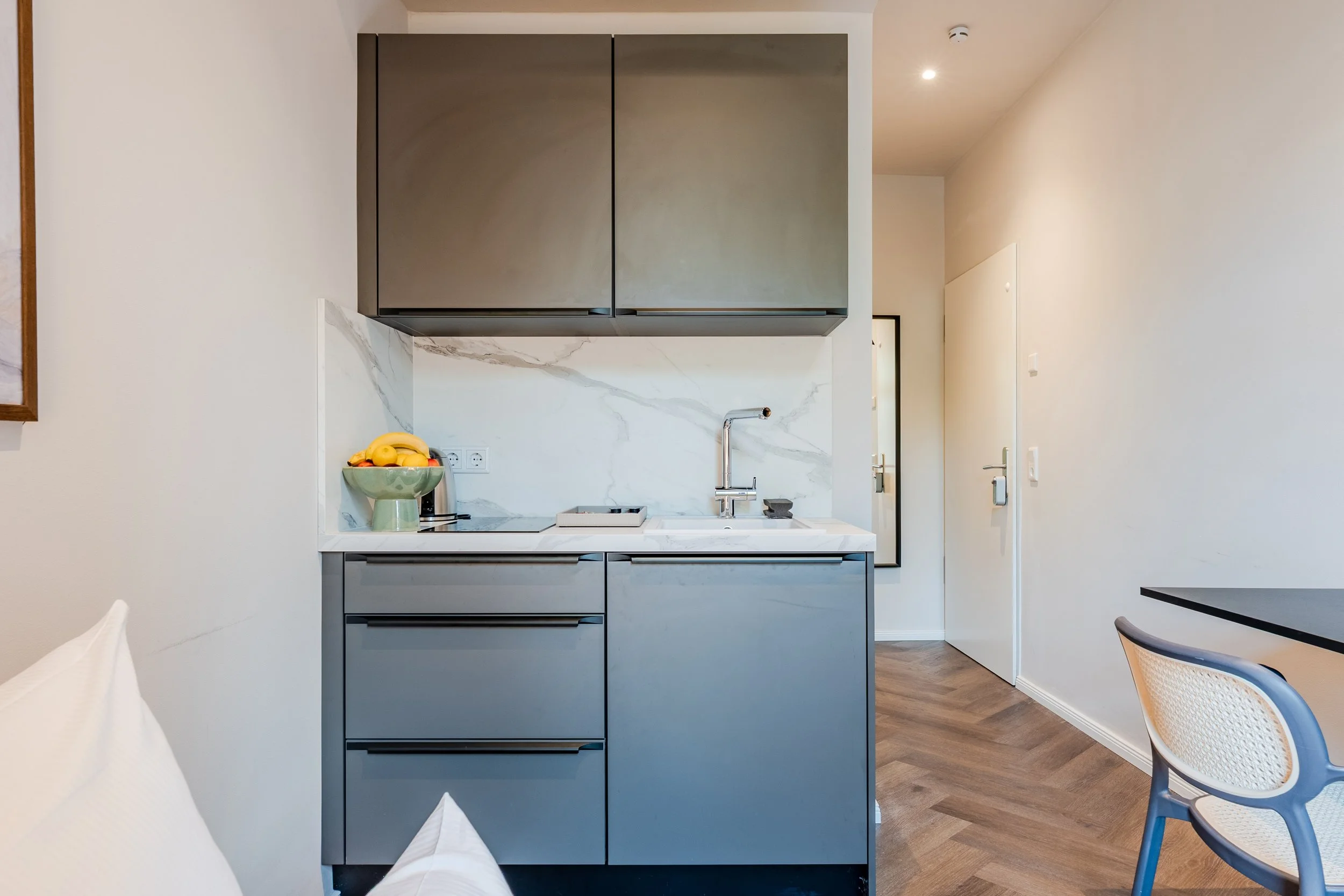 Modern kitchen with gray cabinetry, marble backsplash, stainless steel sink and faucet, fruit bowl, and dining chairs nearby.