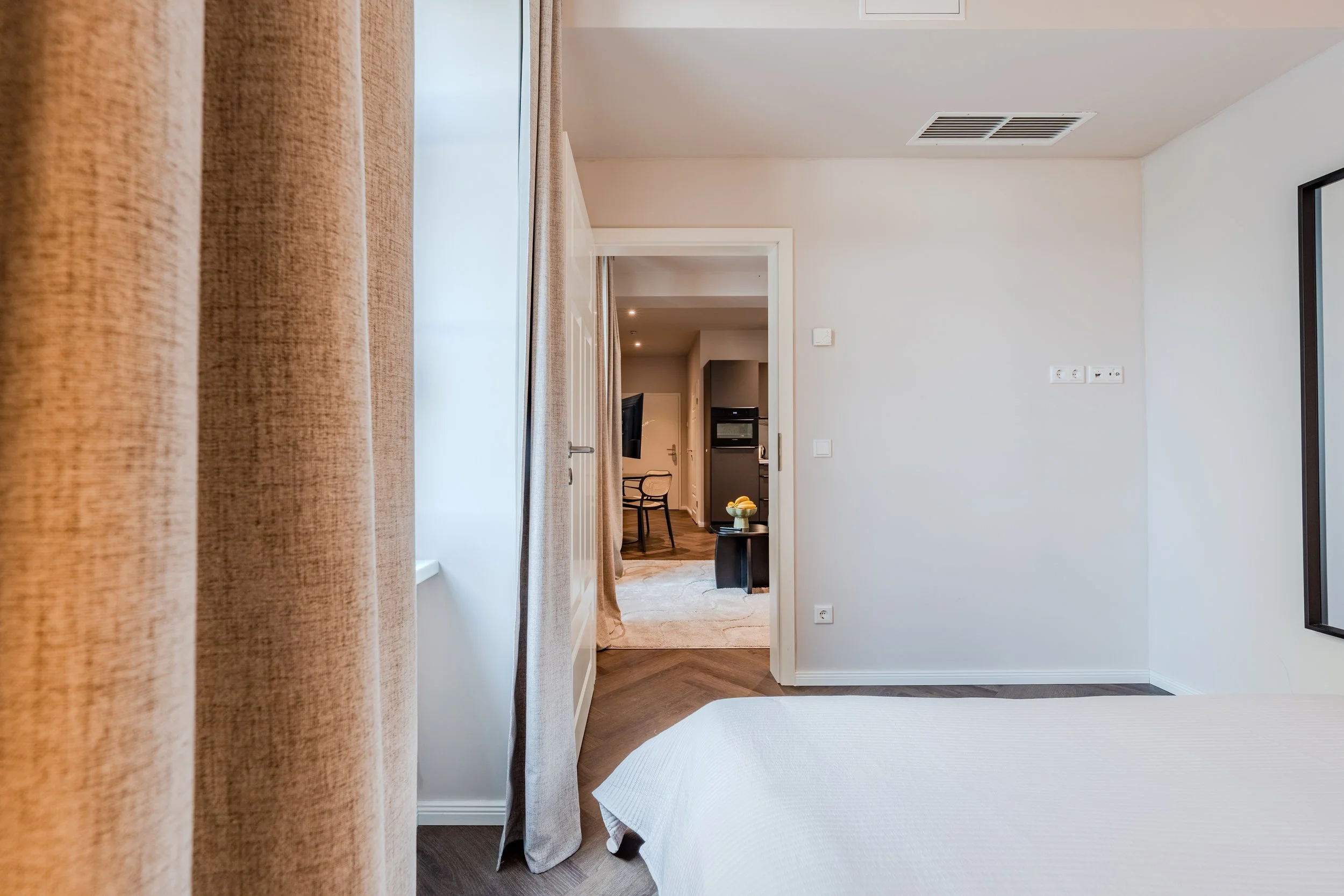 A hotel room with beige curtains, a white bedspread, and a view into a living area with a black chair, coffee table with a bowl of oranges, and a kitchen in the background.