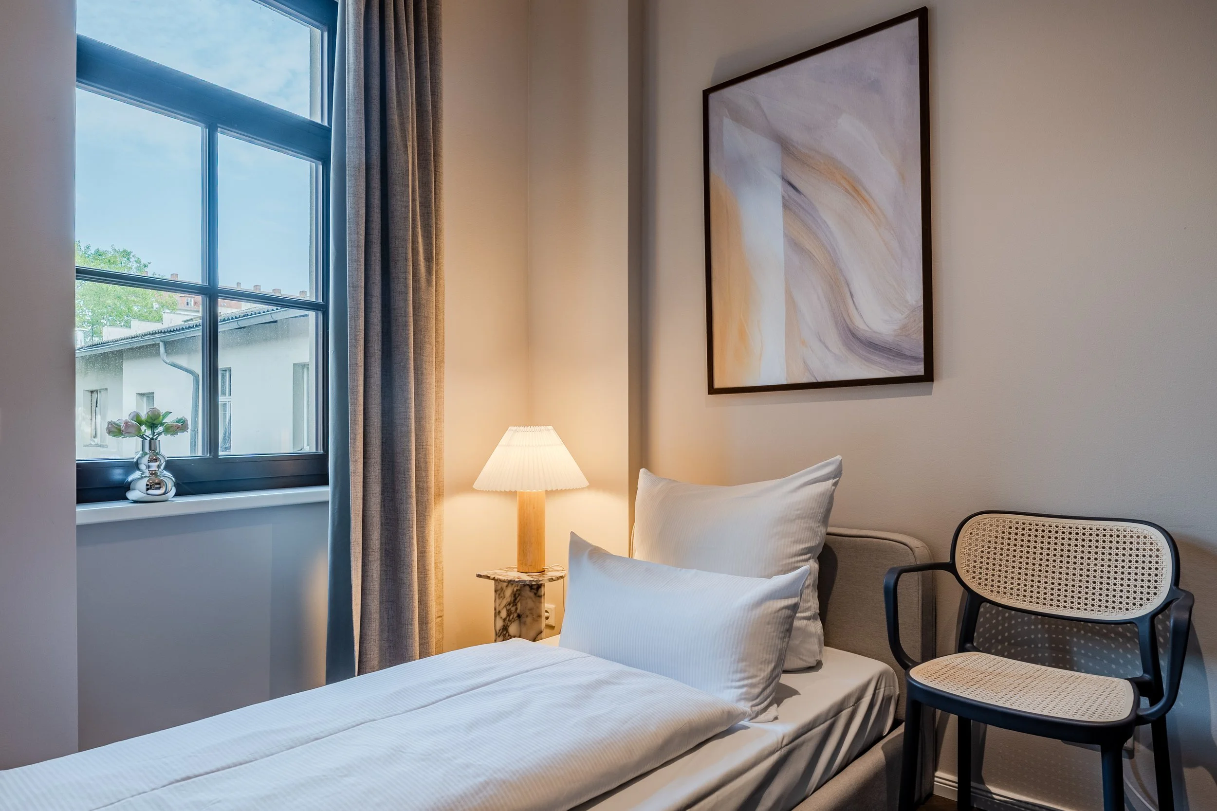 A cozy bedroom corner with a window, a bed with white linens, a table lamp, a piece of abstract art on the wall, and a black chair with a woven cane backrest.