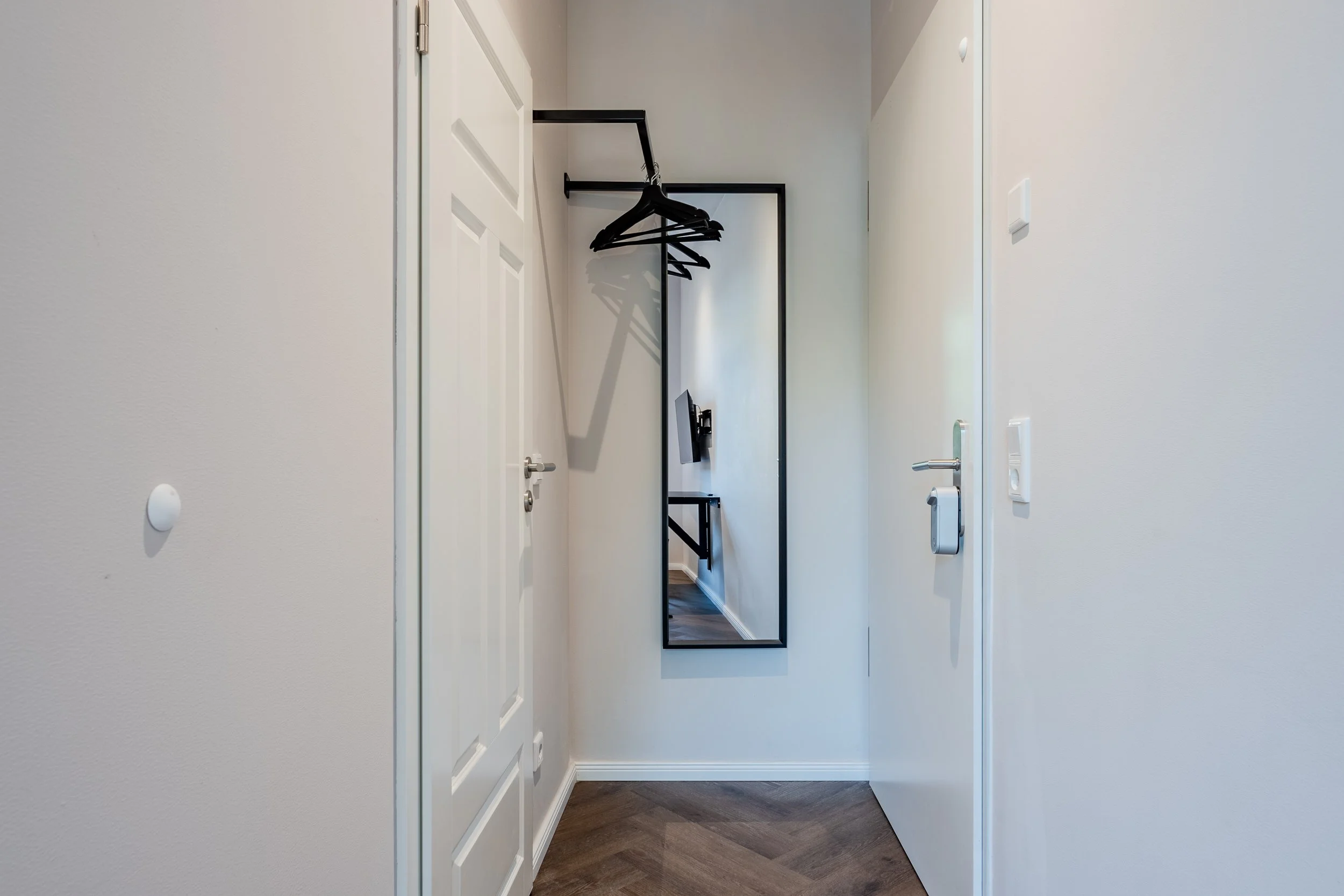 Interior hallway with a white door on the left, a black full-length mirror on the wall, and a small rack with hangers reflecting in the mirror. Cream-colored walls and a dark wooden floor.
