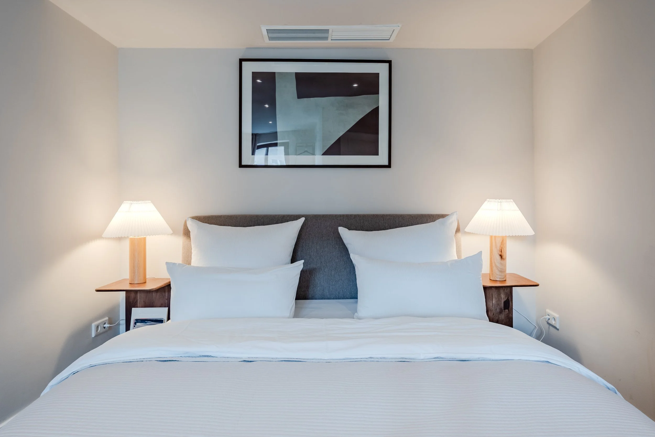 A neatly made bed with white pillows and bedding, flanked by two wooden nightstands with matching table lamps, above which hangs a framed abstract artwork on a light-colored wall in a bedroom.
