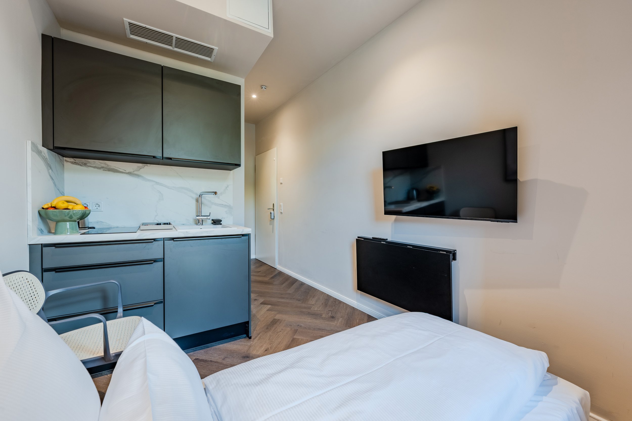 Interior view of a modern hotel room with a kitchenette. The room features a mounted flat-screen TV on the wall, a compact kitchen with black cabinets, a sink, and a small countertop with a bowl of bananas and apples. Part of a bed with white linens and a chair are also visible.