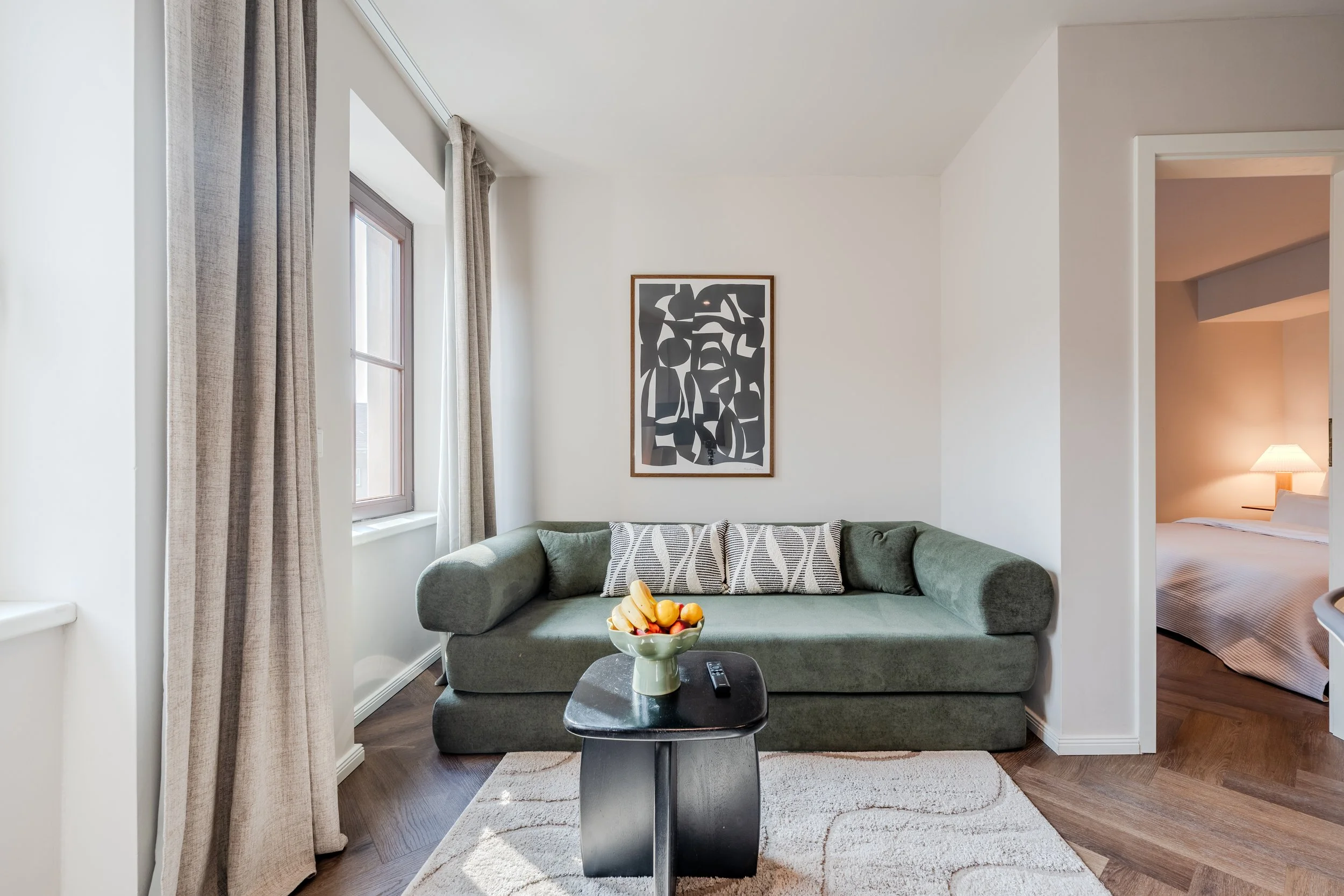 A living room with a green sofa, a black coffee table with a bowl of fruit and two remote controls. Light enters through two windows with beige curtains, and a framed abstract black and white artwork hangs on the wall. A doorway leads to a bedroom with a bed, a nightstand, and a lamp.