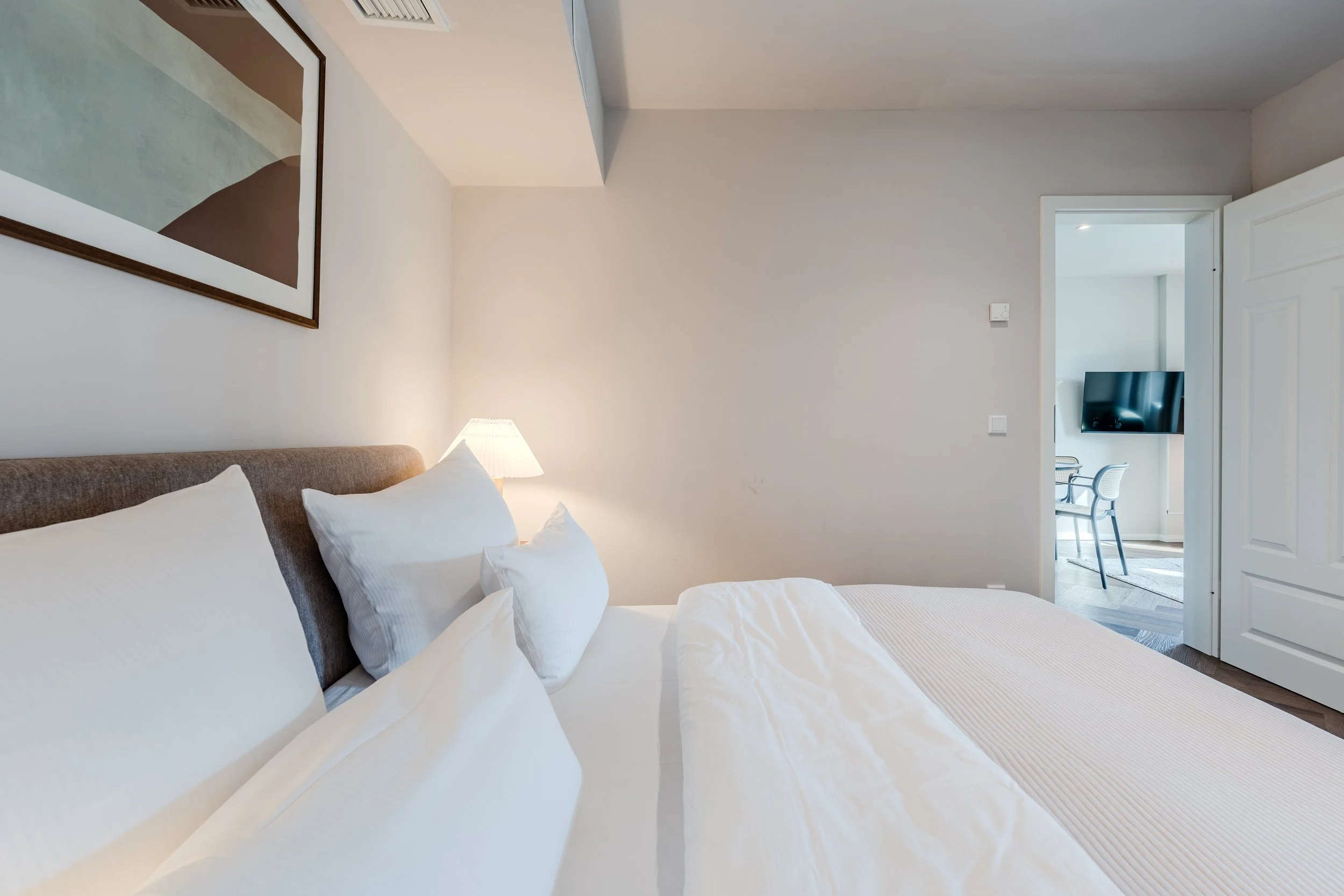 A bedroom with a bed that has white linens and multiple pillows, a bedside table with a lamp, a framed abstract artwork on the wall, and an open door leading to a room with a TV and a chair.