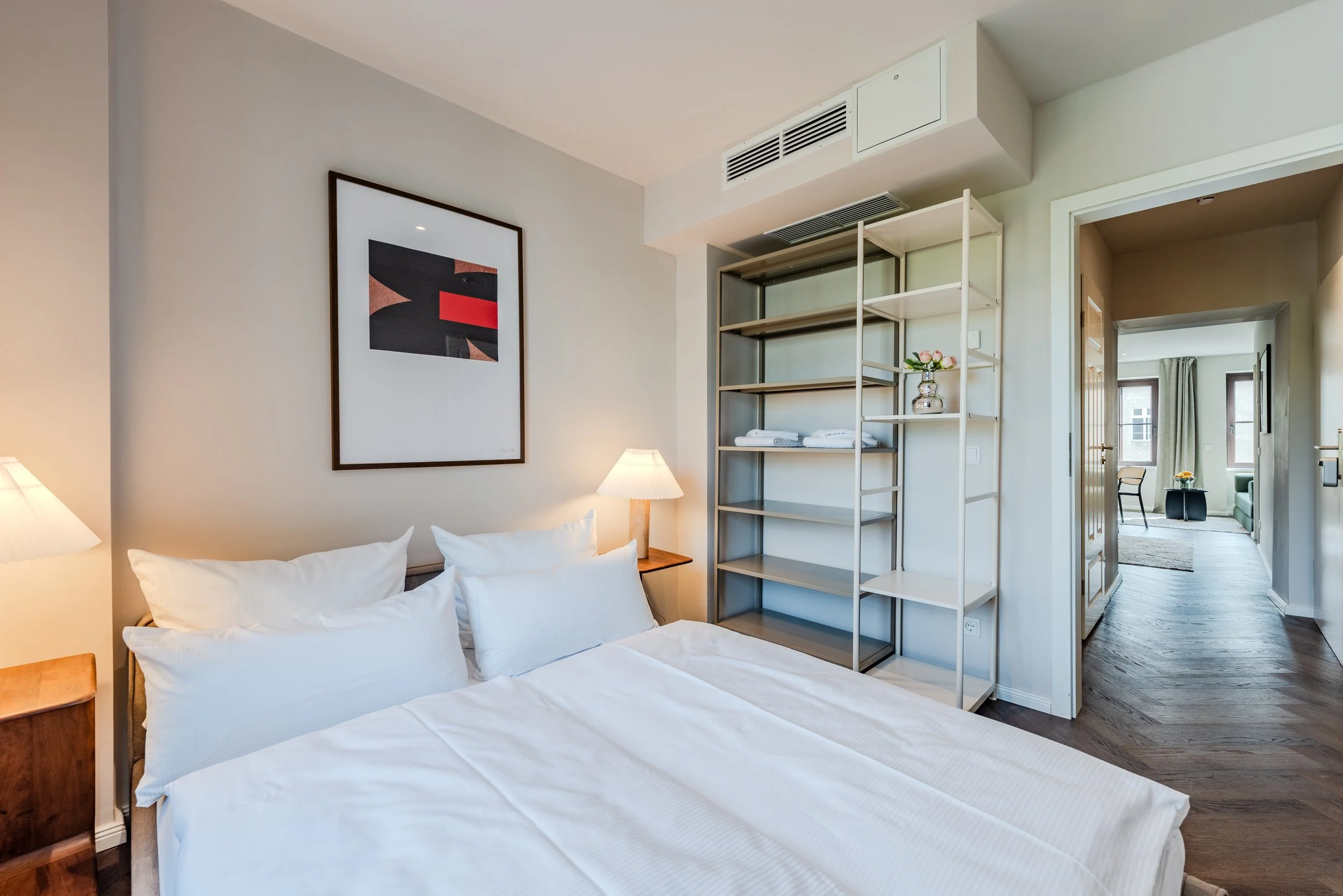 A bedroom with a bed covered in white bedding, a wooden nightstand with a lamp, abstract artwork on the wall, a shelving unit with towels and a flower vase, visible doorway leading to another room with seating and large windows.