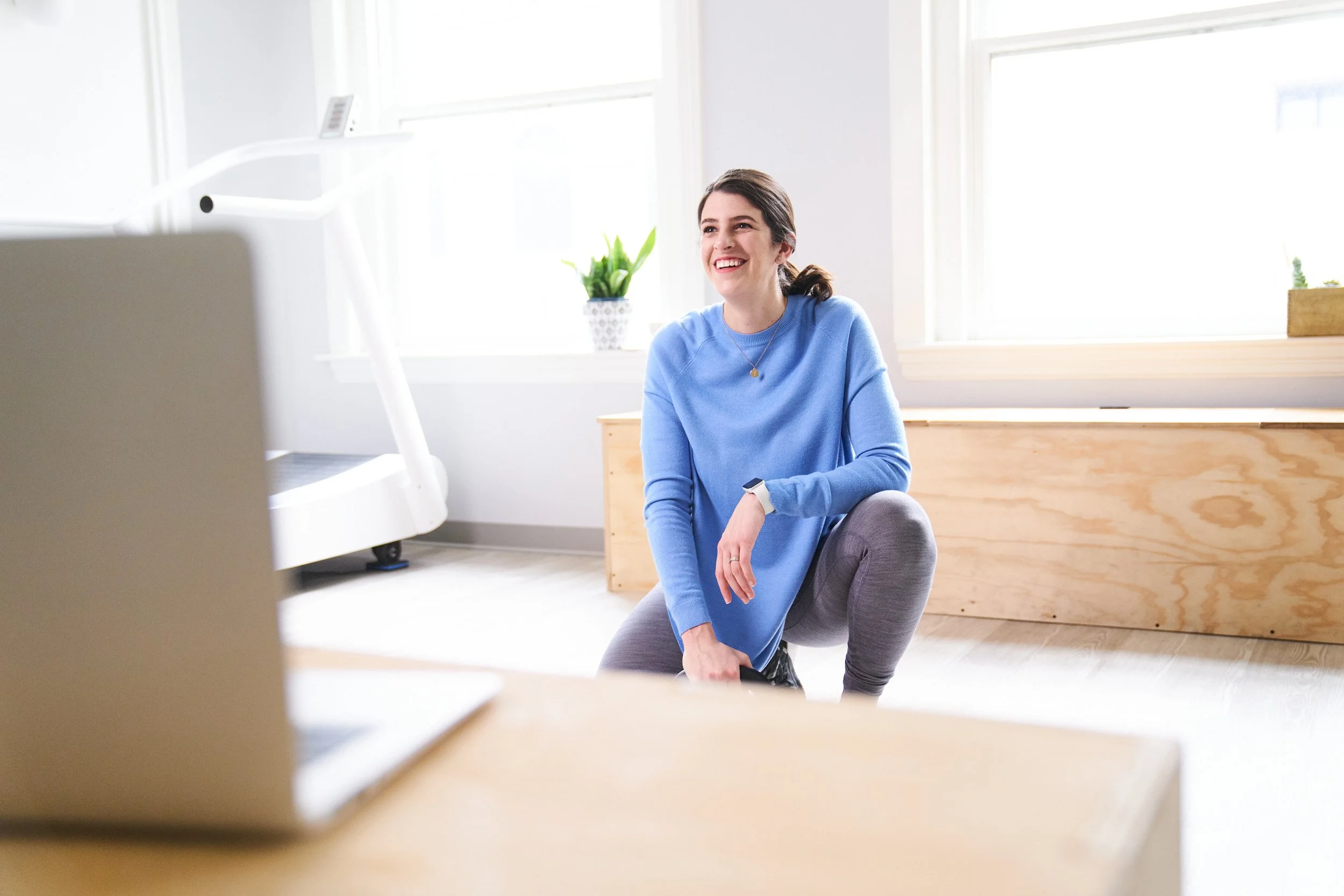 A woman smiling and sitting on a yoga ball in a bright, modern room with large windows and minimal decor.