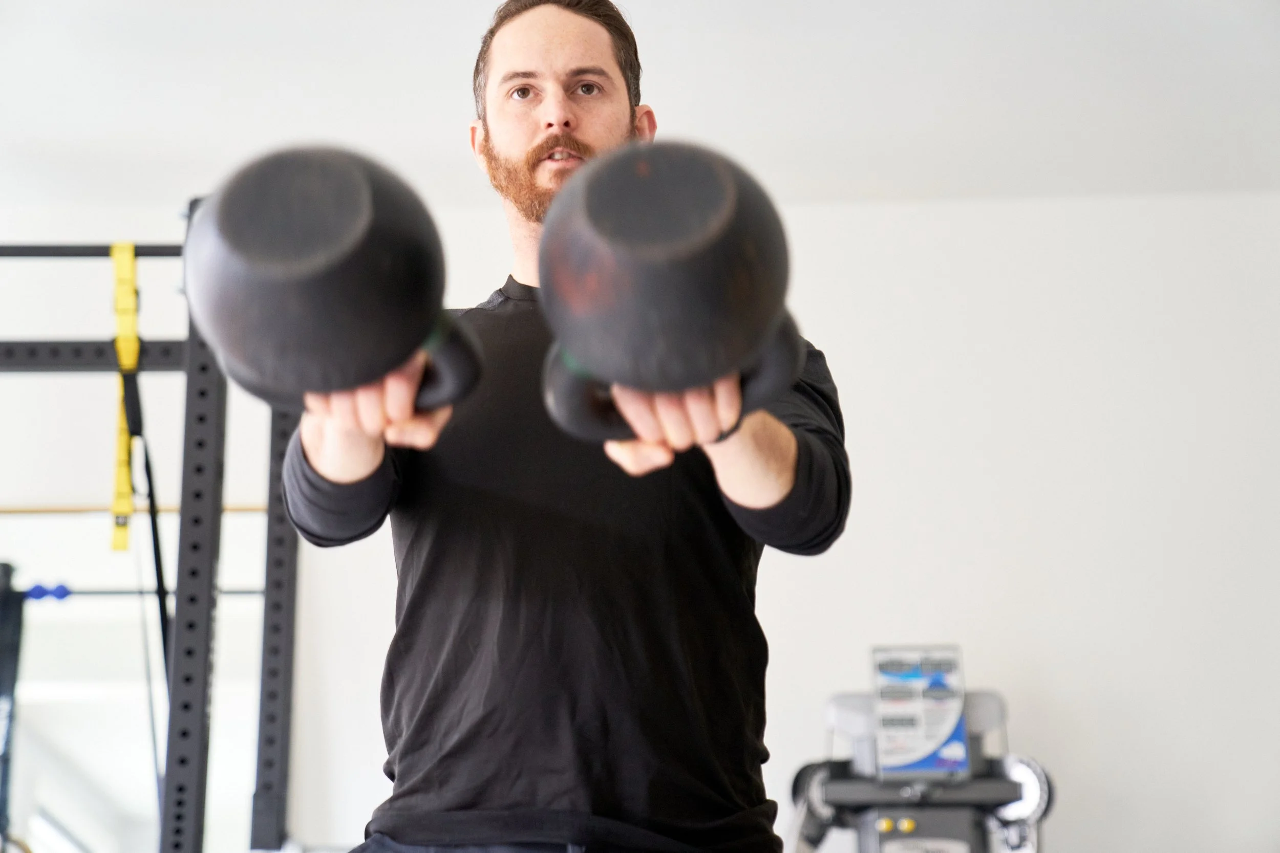 A man with a beard lifts two black kettlebells during a workout in a gym setting.