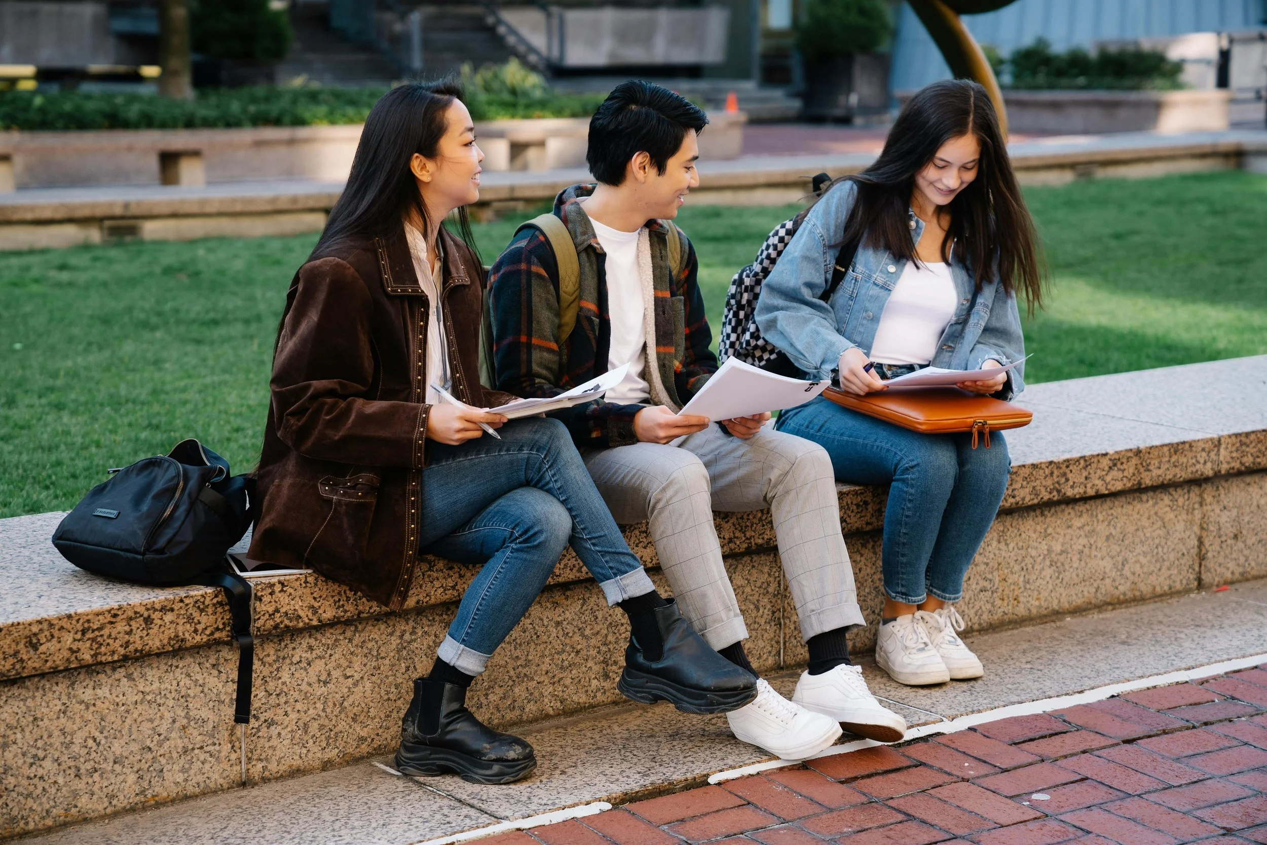Three college students smiling and comparing notes on paper are sitting outside on a raised ledge surrounding a grassy area