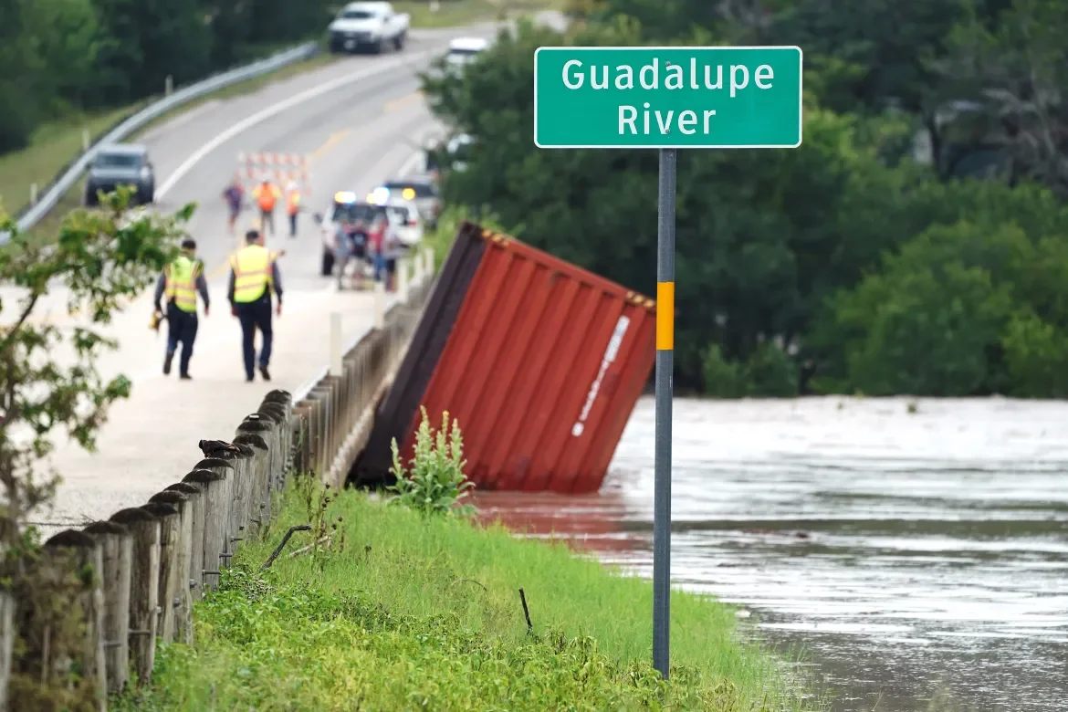 Guadalupe River floodwater beside a highway, with a red shipping container tipped against a roadside fence and emergency crews walking the road