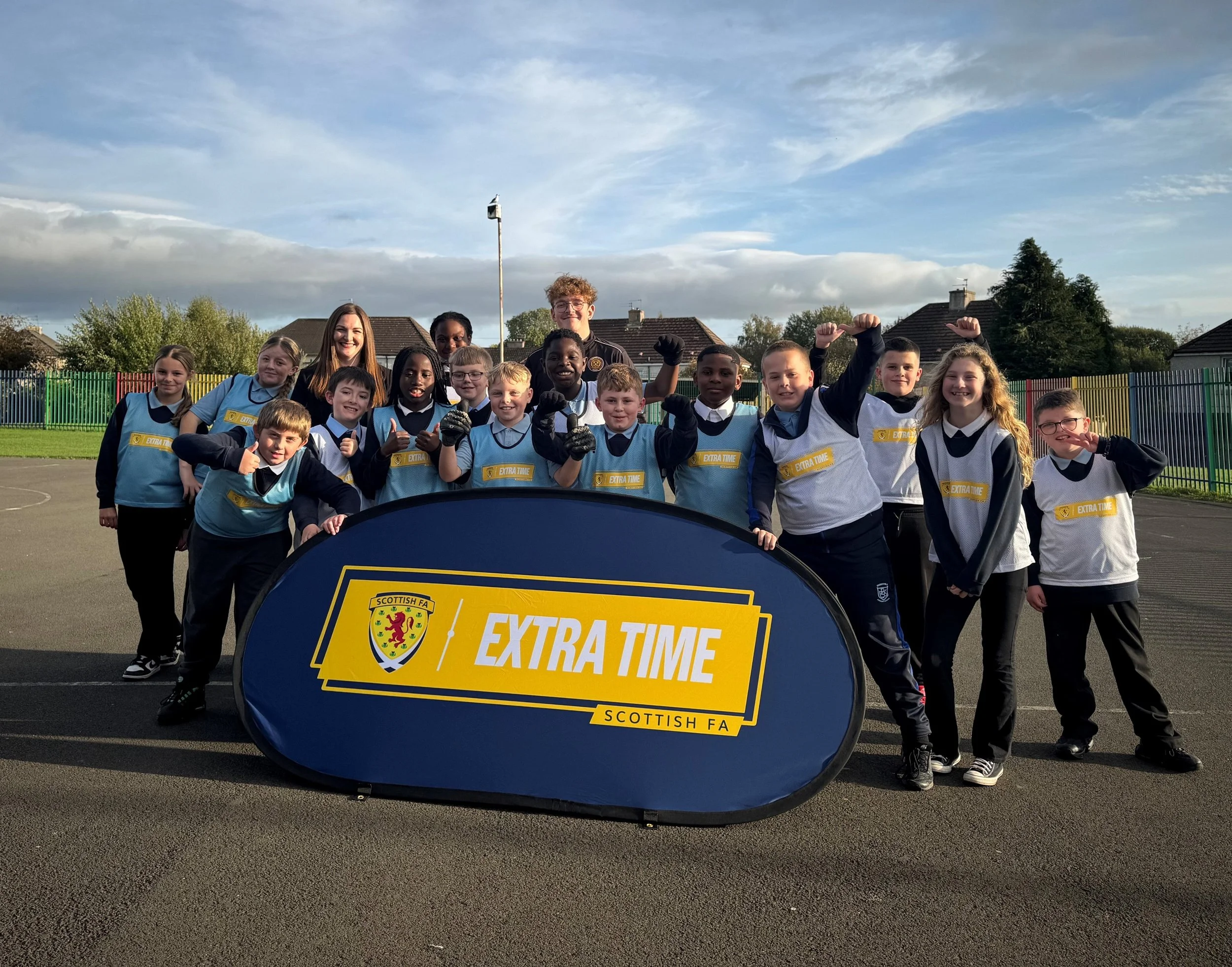 A group of children and two adults standing outdoors on a sports field, posing for a photo behind a sign that says "Extra Time" with the Scottish FA logo. Some children are wearing sports uniforms and smiling, with a few raising their fists. The sky is partly cloudy with blue patches, and there are trees and houses in the background.