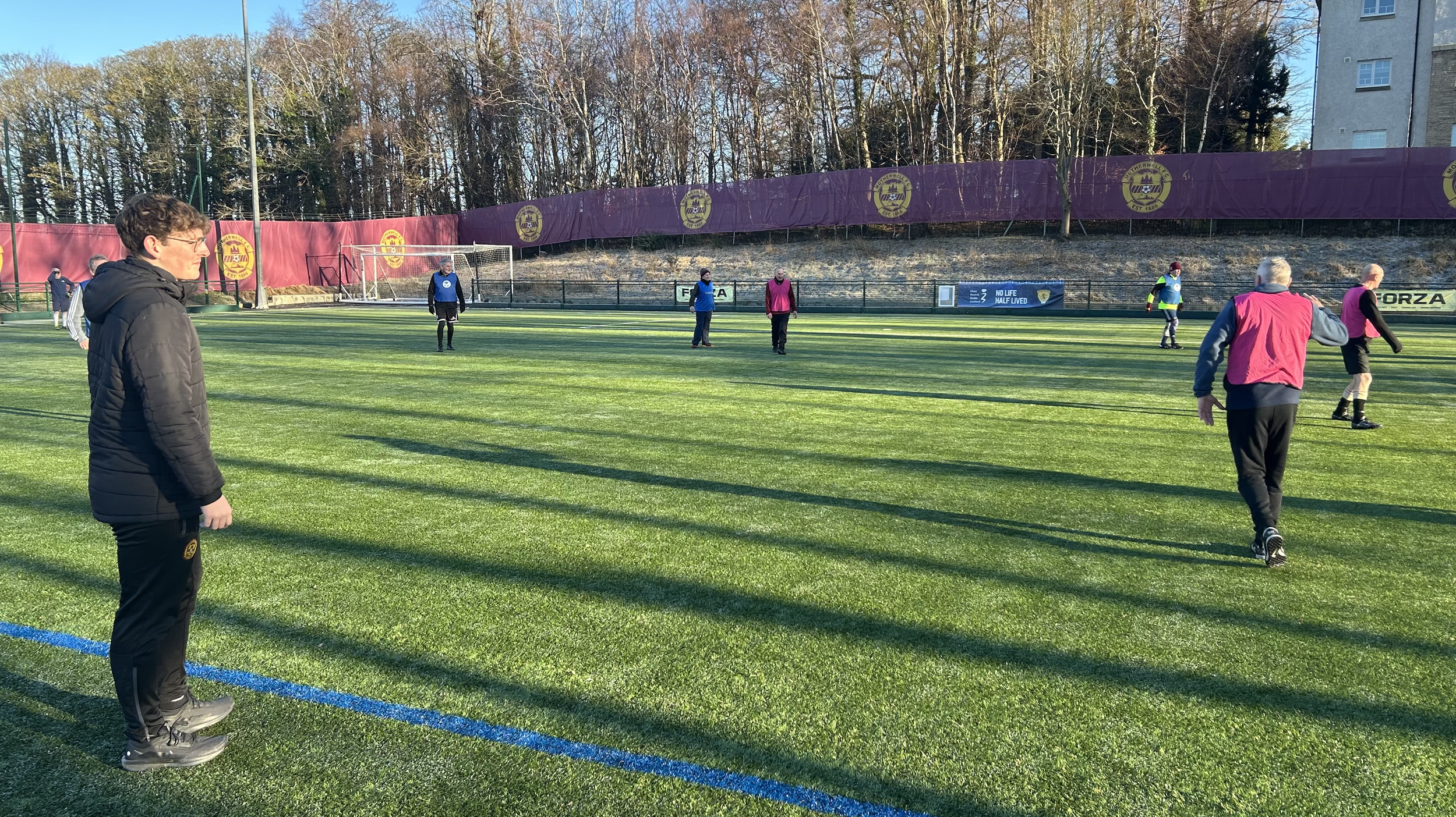 People playing soccer on a green field with purple flags bearing a club emblem, trees in the background, and some individuals in pink and blue training vests.
