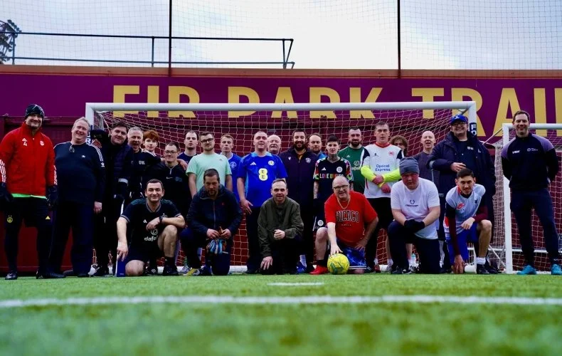 Group of men and boys posing on a soccer field in front of a goal with a sign that reads 'FIR PARK TRAIL' in the background.