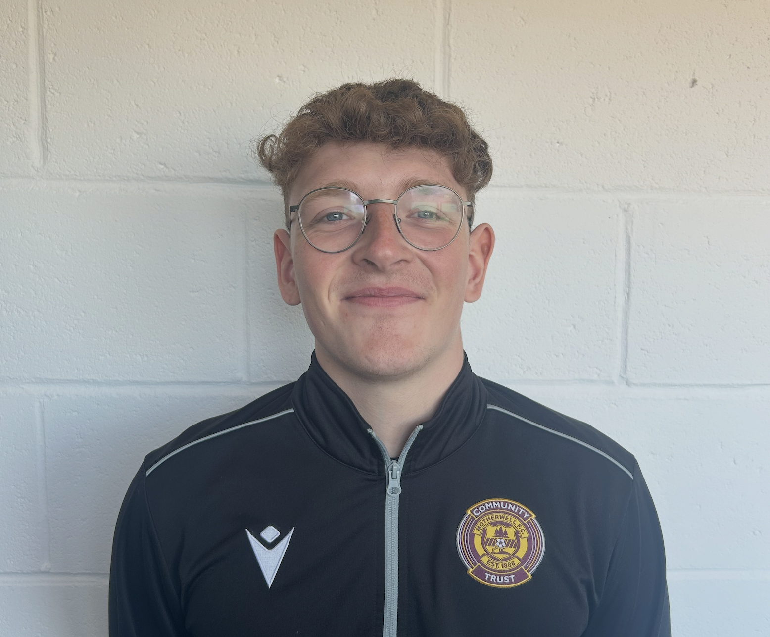 A young man with curly brown hair and glasses, wearing a black sports jacket with a crest on the left chest that reads 'Motherwell F.C. Trust,' stands against a white cinder block wall.