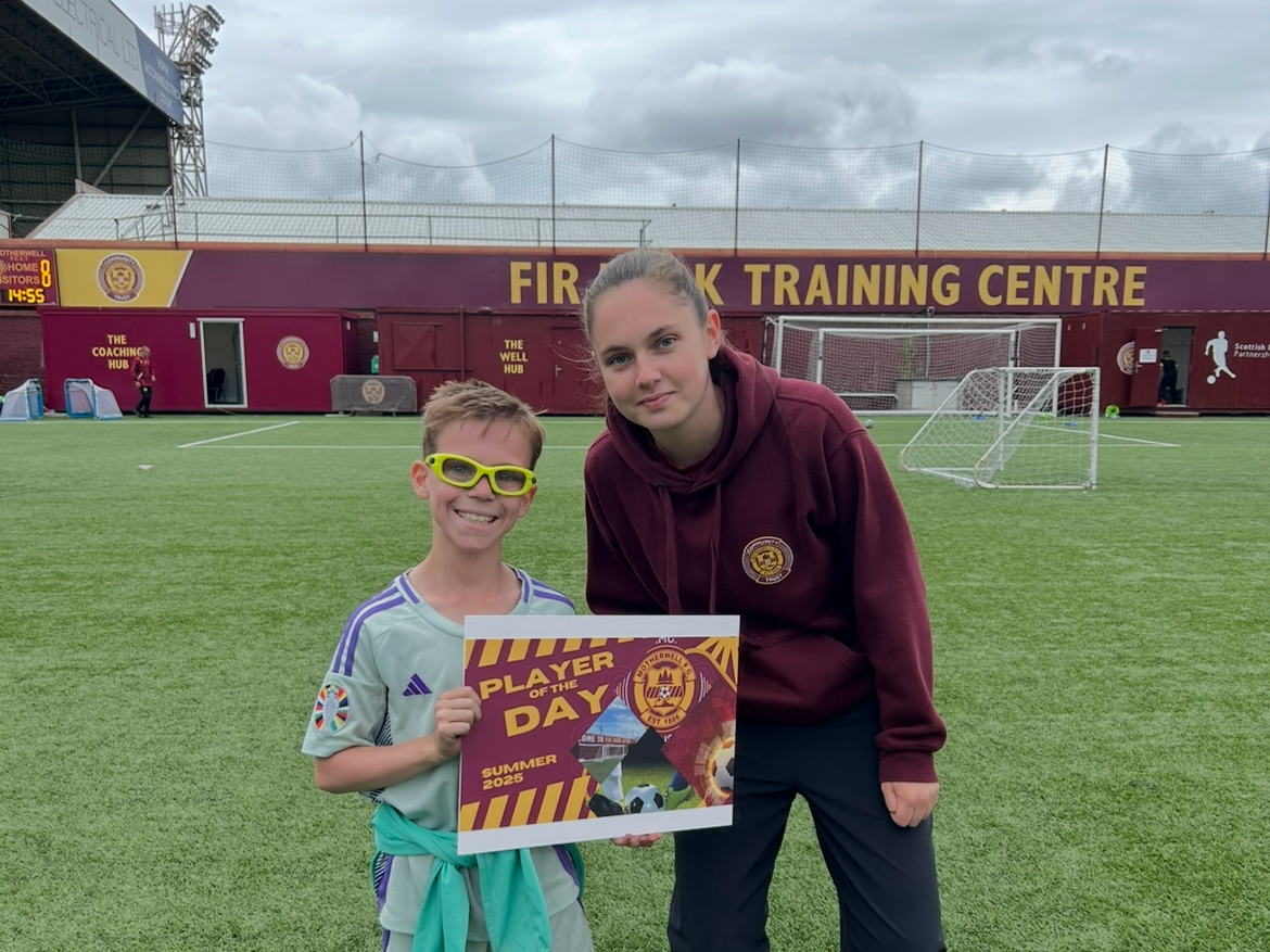 A young boy and a woman at a soccer field holding a sign that says 'Player of the Day Summer 2025'. The boy is smiling and wearing yellow glasses and a soccer jersey, while the woman is wearing a maroon hoodie with a crest. The soccer field and training center are visible in the background.