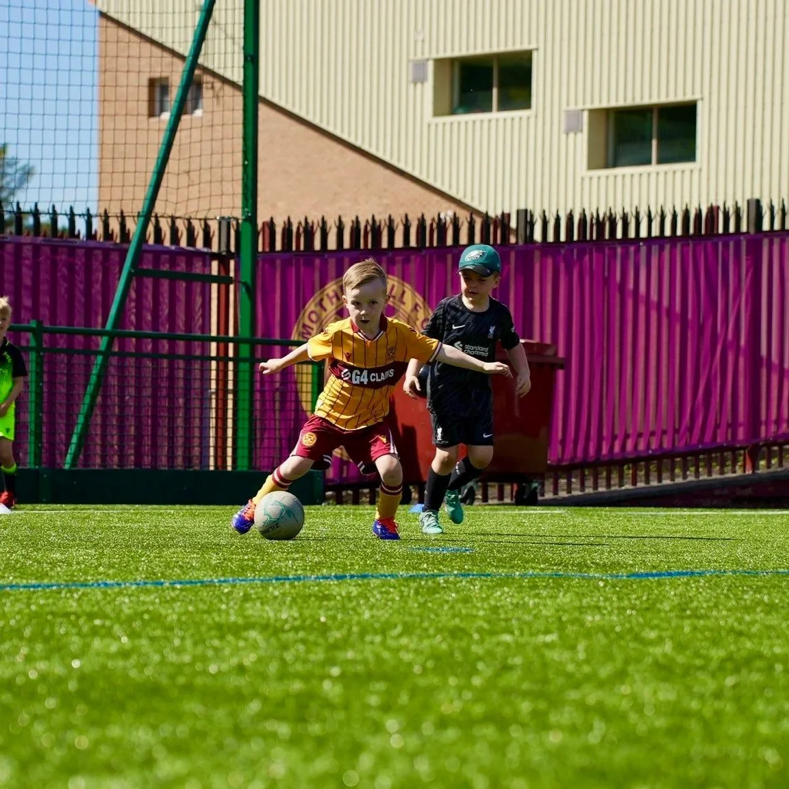 Children playing football on a green field with a colorful fence and buildings in the background.