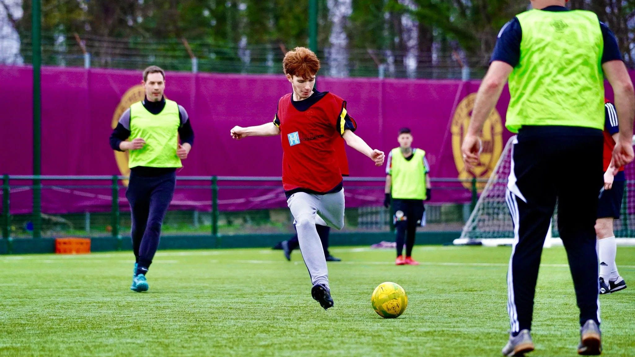 People playing soccer on a grassy field, with one young man kicking a yellow soccer ball while others watch.