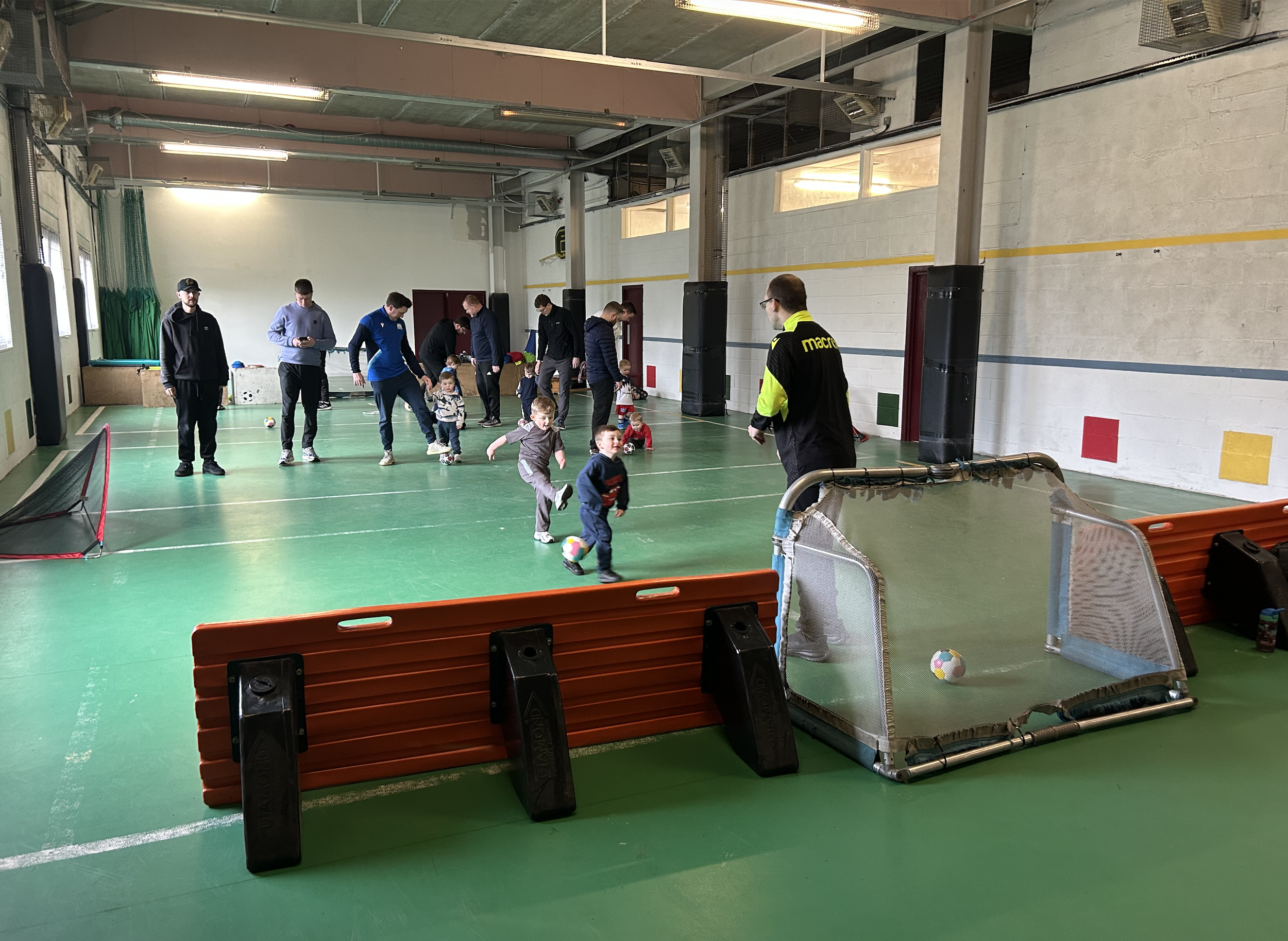 Indoor gymnasium with children and adults playing with footballs during a sports activity class. Several footballs are scattered on a green floor, with some running or sitting children and adults standing and observing.