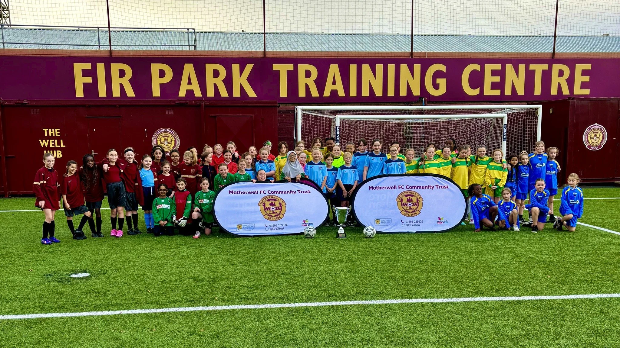 Group of children and young players in sports uniforms posing on a soccer field in front of a large sign that reads 'FIR PARK TRAINING CENTRE.' There are two banners with the logo of Motherwell FC Community Trust and several sports trophies in front of the group.