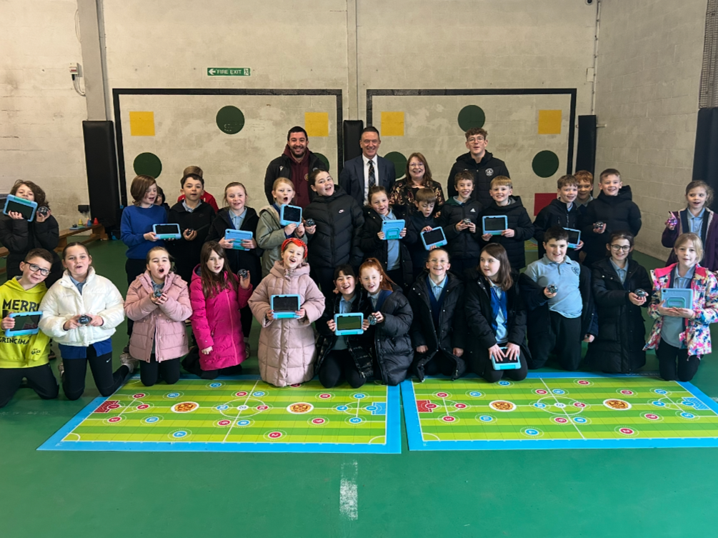 Group of children and adults posing in a gymnasium with children holding tablets, some kneeling in front and some standing behind, on a bright green field with colorful game markings.
