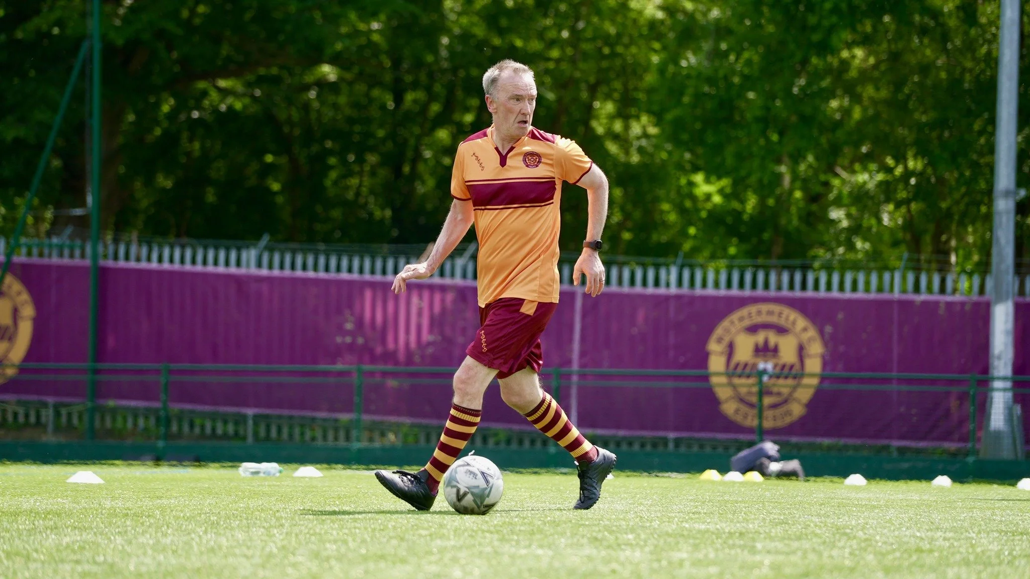 An elderly man wearing a maroon and gold sports uniform practicing soccer on a field, with a soccer ball, cones, and training equipment around him.