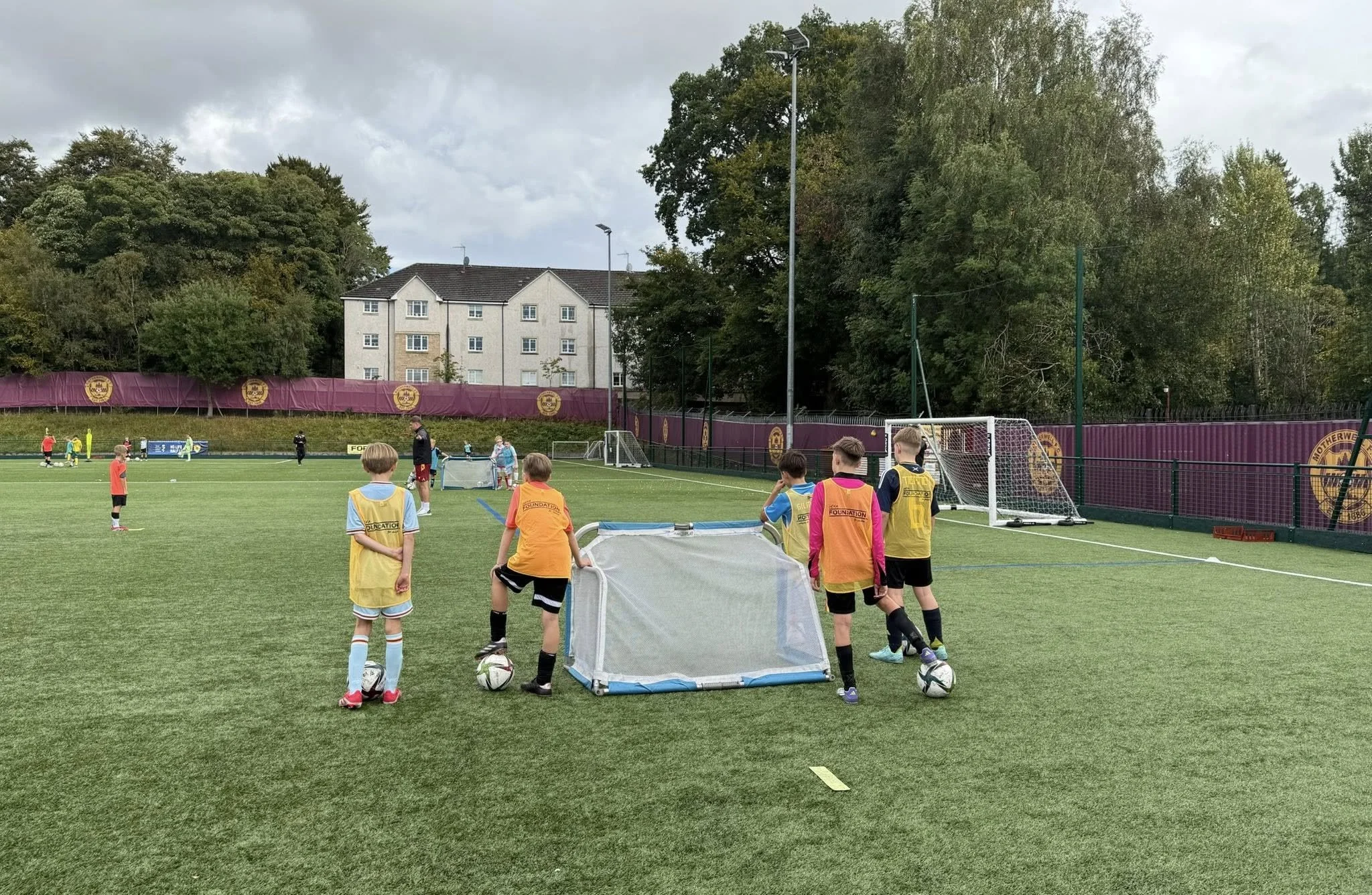 Children participating in a youth soccer practice on a field, with coaches, small goals, and a purple fence with team logos, surrounded by trees and residential buildings under cloudy skies.