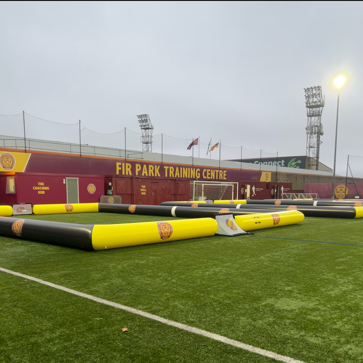 An outdoor training area at Fir Park Training Centre featuring inflatable goalposts and barriers on a grassy field, with a building displaying the name and logos in the background.